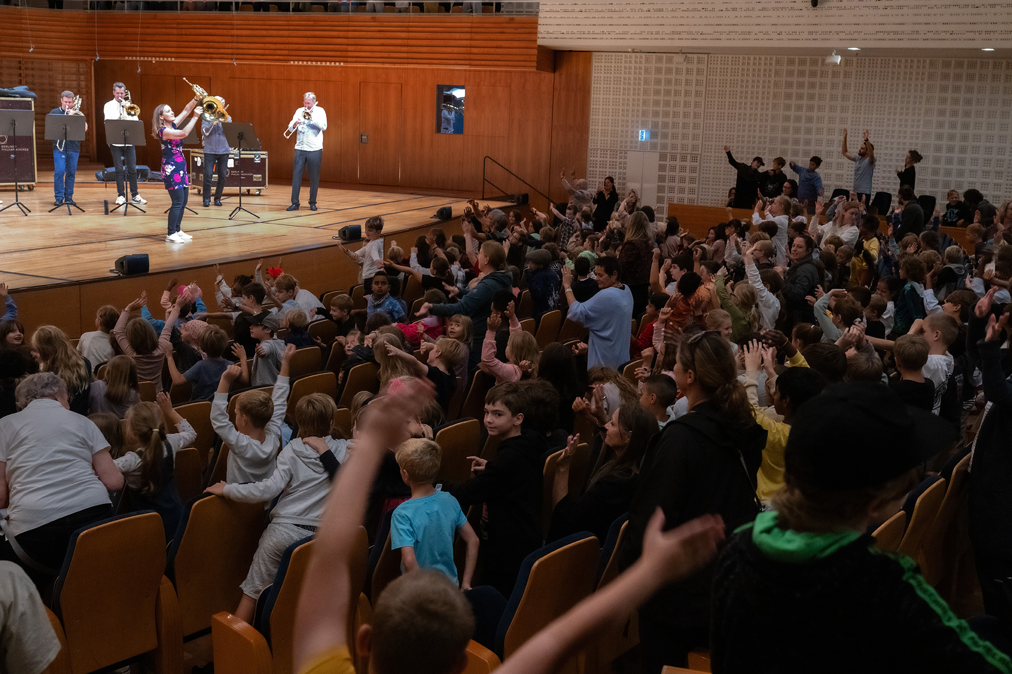 Kinder im Publikum mit hochgereckten Armen, auf der Bühne Musiker mit Blechblasinstrumenten