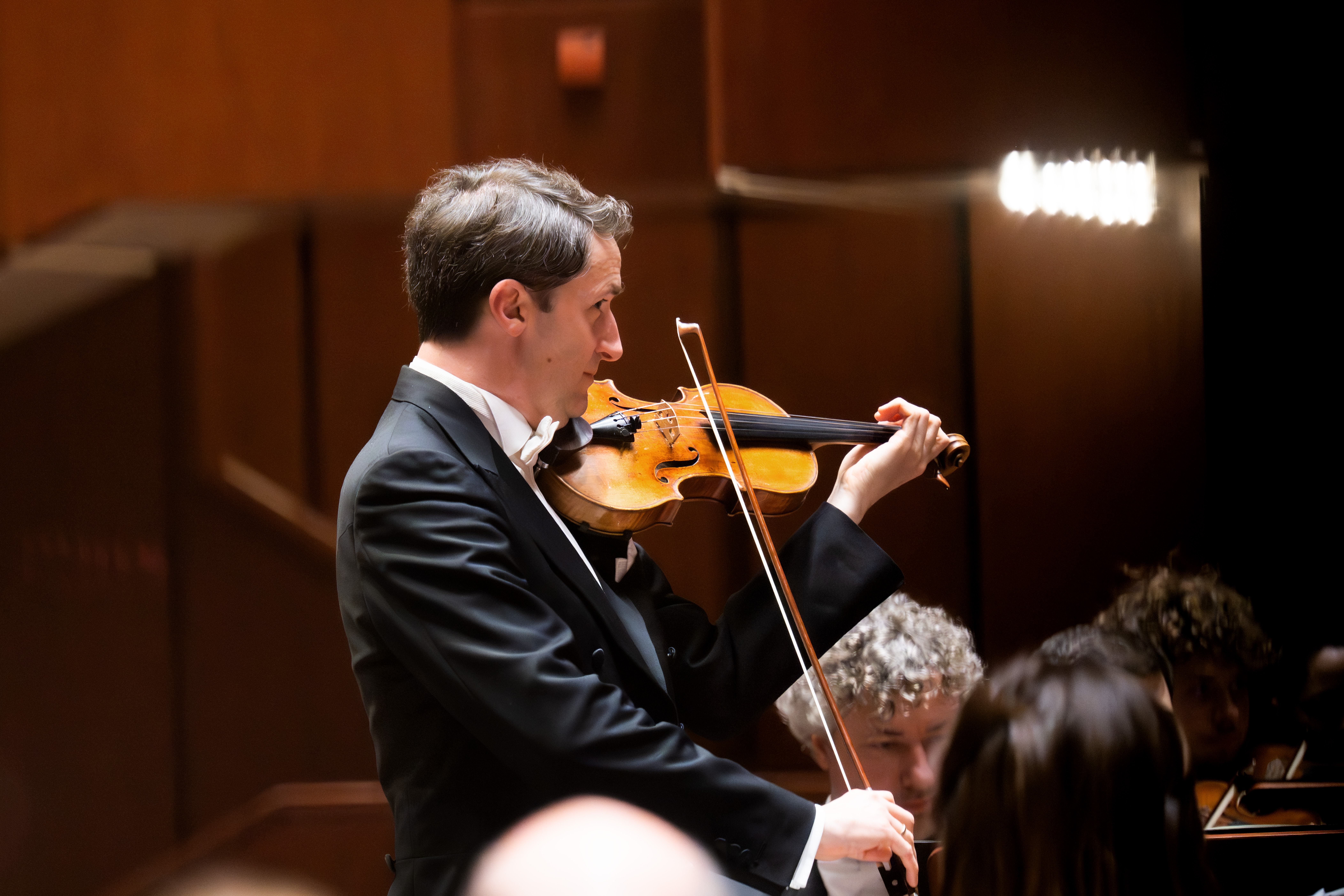 Noah Bendix-Balgley in a black tuxedo plays the violin on stage with an orchestra, focused and illuminated by warm stage lighting. Other musicians and a wooden background are visible behind him.
