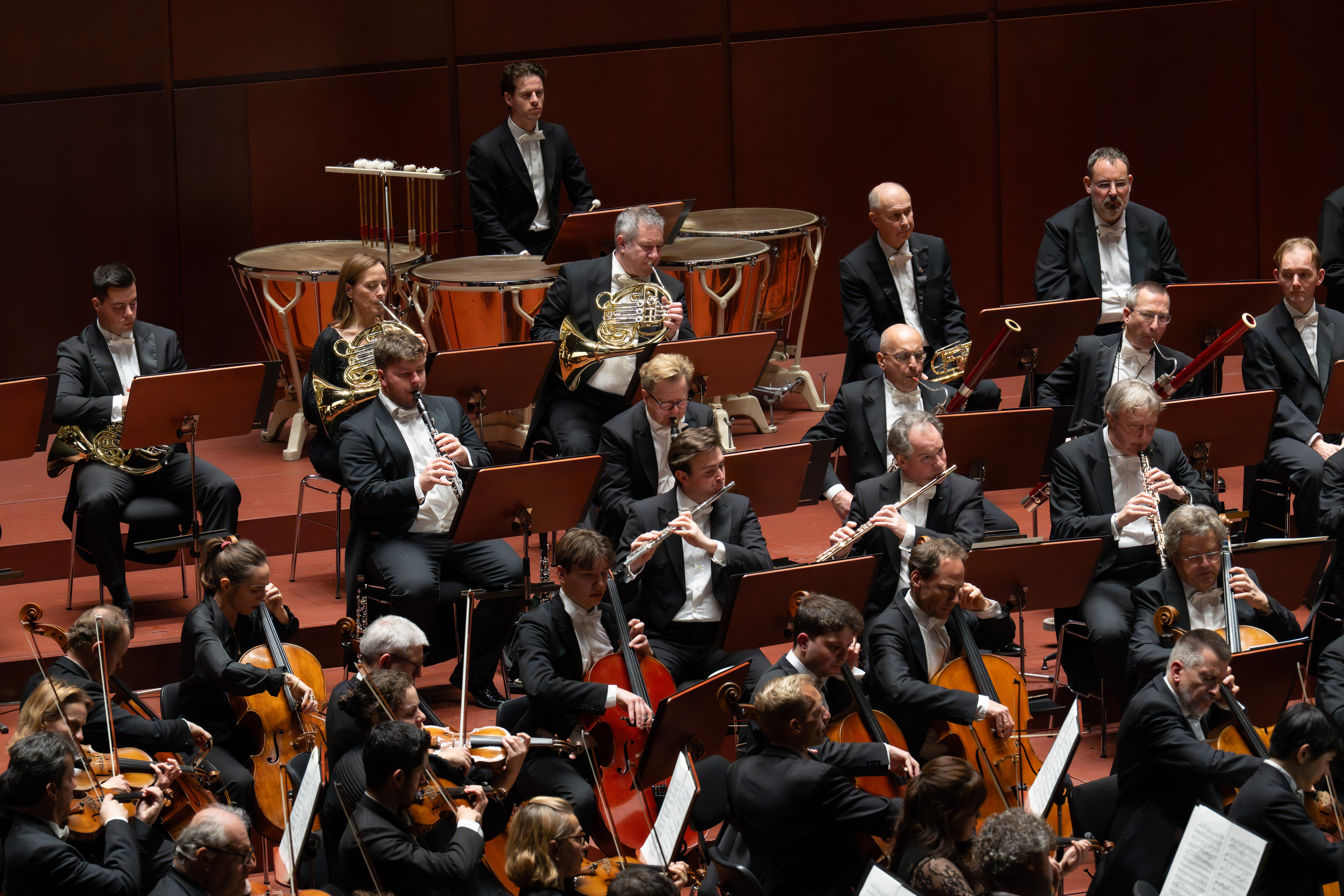 On stage, the Berliner Philharmoniker perform — musicians on woodwinds, brass, and strings, all dressed in formal black attire.