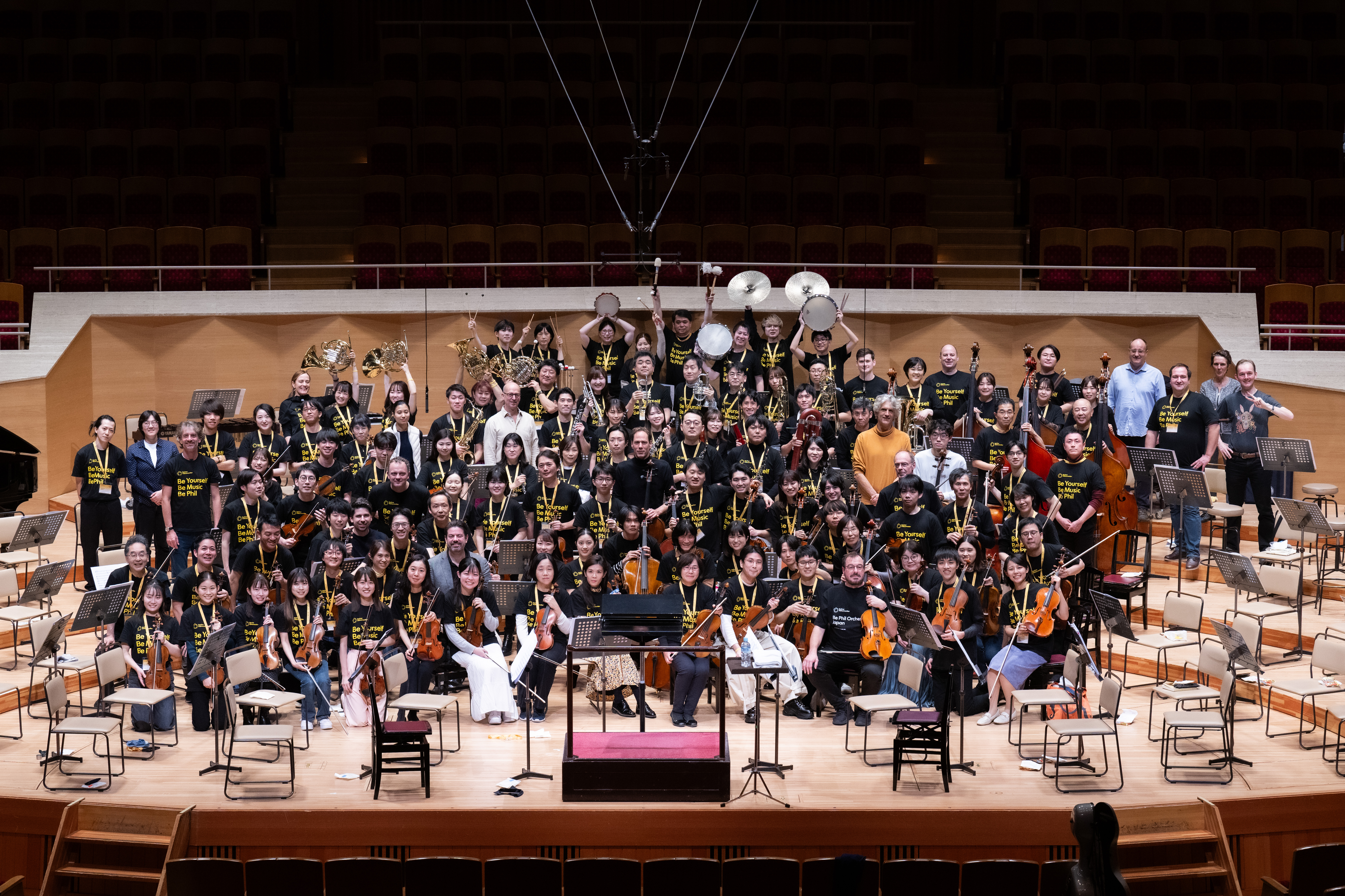 Ein großes Sinfonieorchester posiert auf der Bühne für ein Gruppenfoto. Die Musiker halten verschiedene Instrumente wie Geigen, Hörner und Schlagzeug und tragen alle passende schwarze Hemden in einem Konzertsaal.