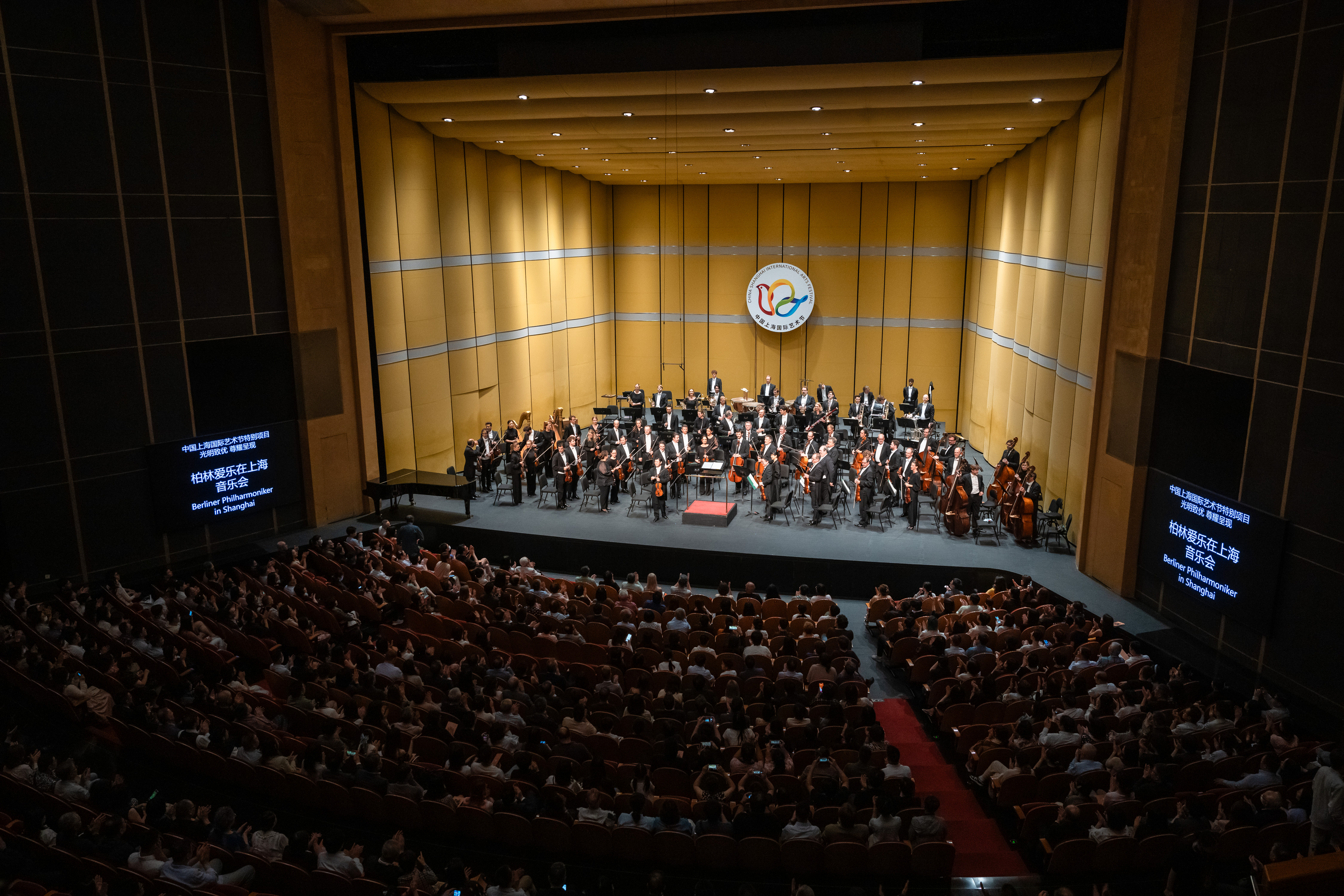 Die Berliner Philharmoniker spielt auf einer großen, hell erleuchteten Bühne mit Musikern, die in Reihen sitzen. Das Publikum füllt das Theater und schaut aufmerksam zu. Zwei Bildschirme zeigen auf beiden Seiten der Bühne Text in chinesischer Sprache an.