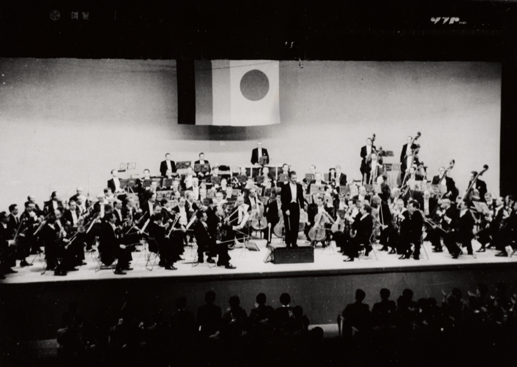 Ein großes Orchester spielt auf der Bühne unter einer japanischen Flagge. Der Dirigent steht in der Mitte und die Musiker sitzen in Reihen vor dem sichtbaren Publikum. Das Bild ist in schwarz-weiß gehalten.