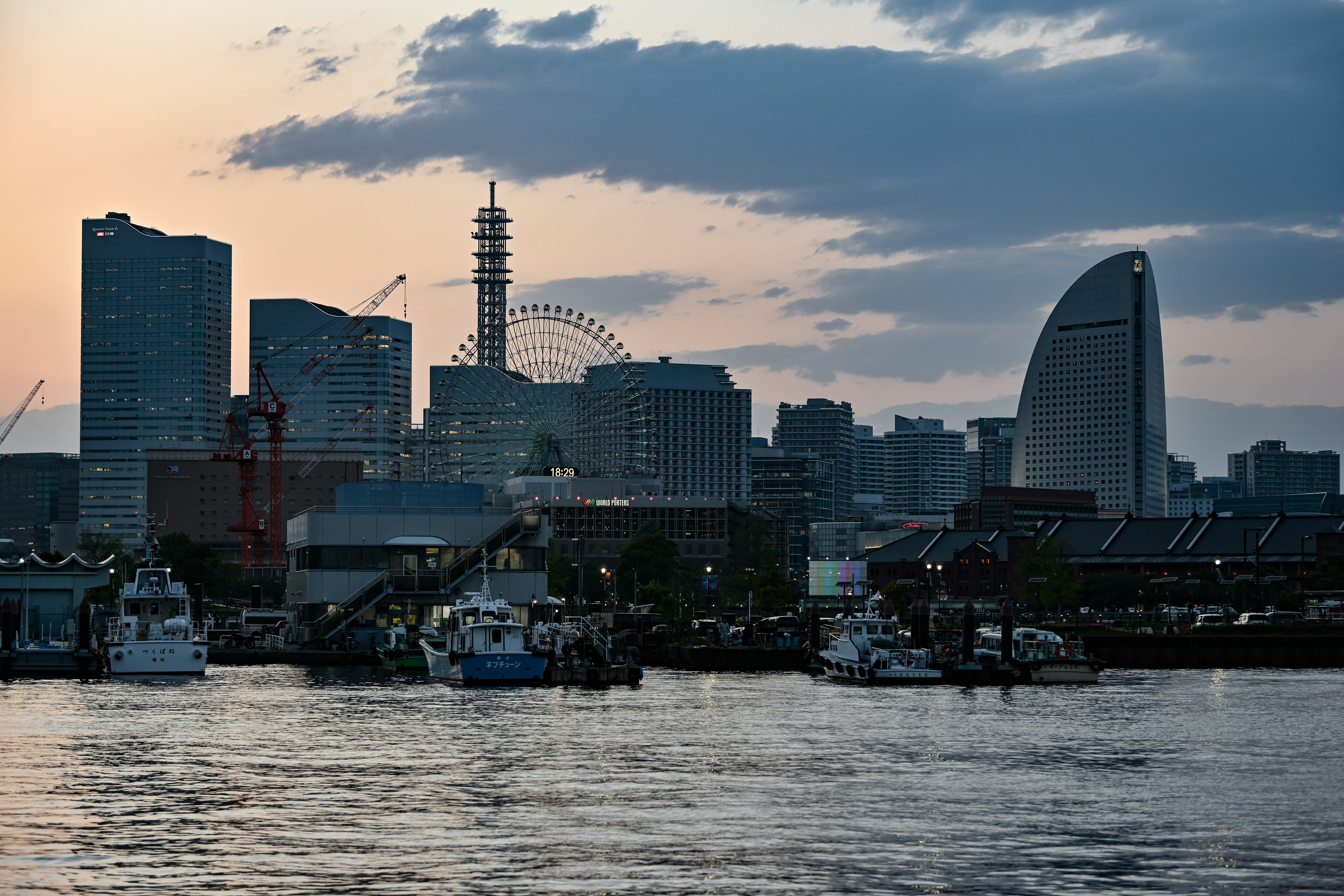 Die Skyline der Stadt in der Abenddämmerung mit modernen Hochhäusern, einem Riesenrad und an der Uferpromenade angedockten Booten, die sich im ruhigen Wasser unter einem teilweise bewölkten Himmel spiegeln.
