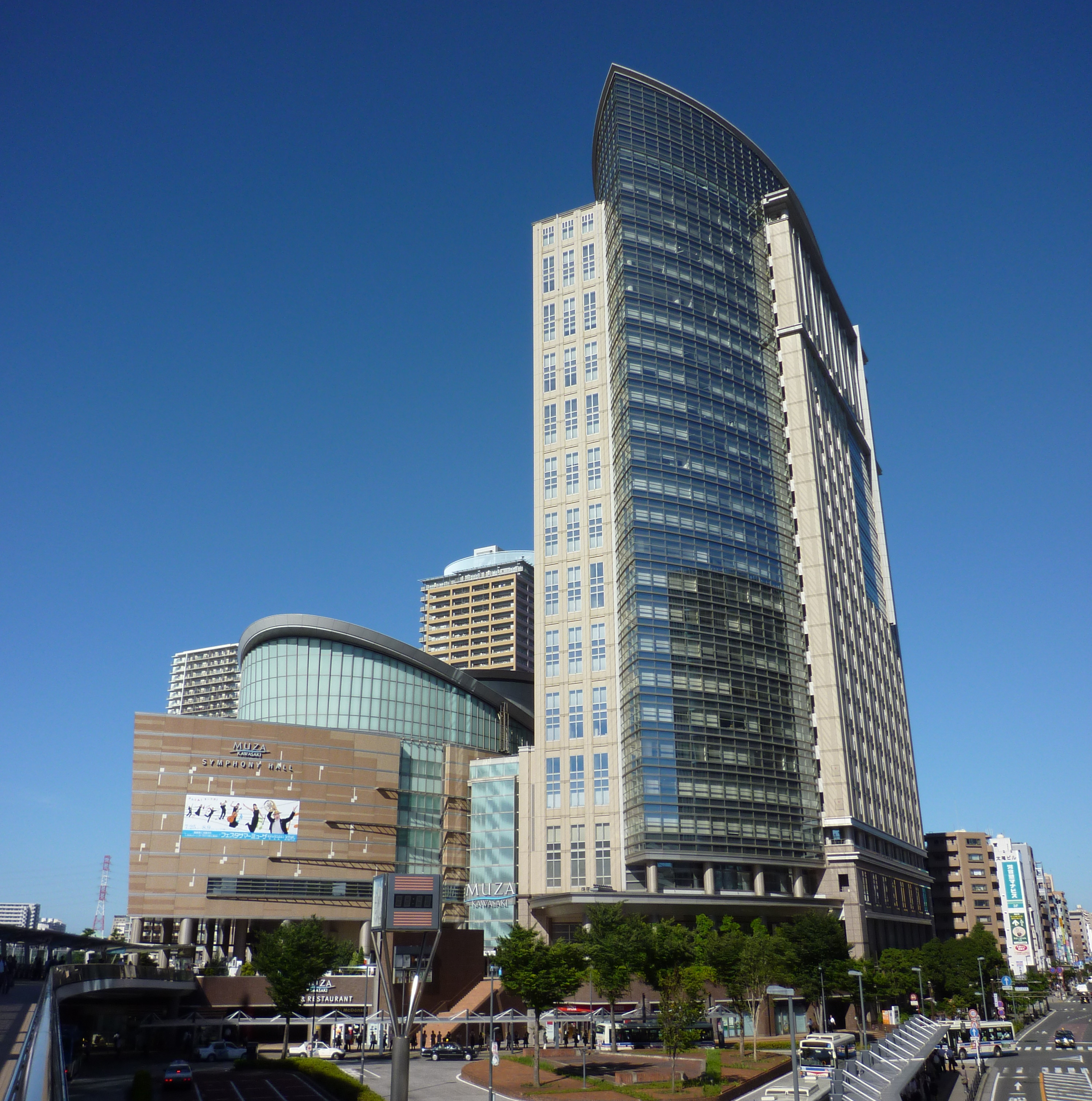 Ein hohes modernes Gebäude mit einer geschwungenen Glasfassade steht vor einem strahlend blauen Himmel, umgeben von anderen Gebäuden und Grünflächen auf Straßenniveau.