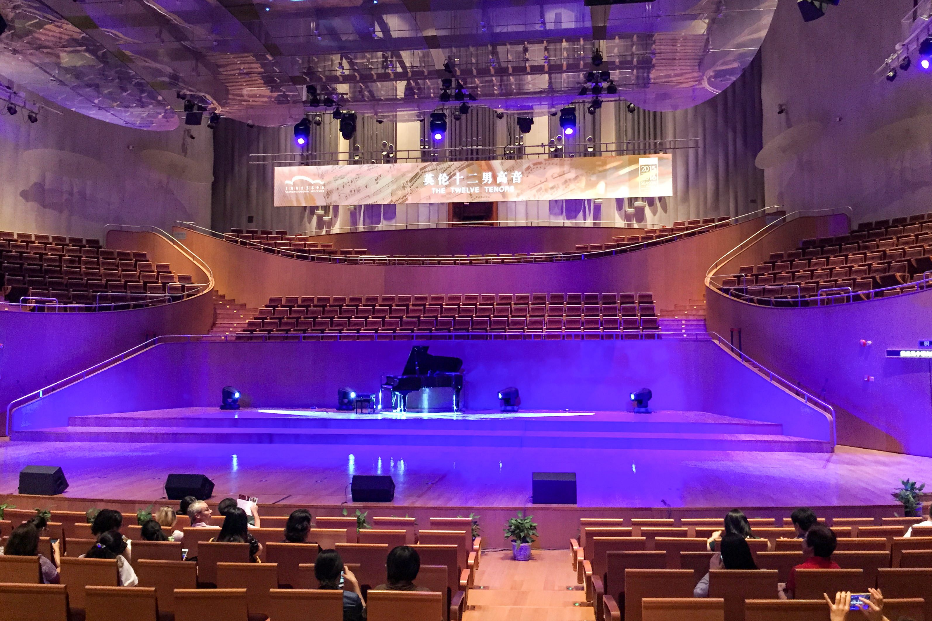 Ein moderner Konzertsaal mit einem Flügel auf der Bühne, leeren Sitzen auf der Bühne und in den oberen Rängen und einigen Zuschauern in den unteren Reihen bei violetter Beleuchtung.