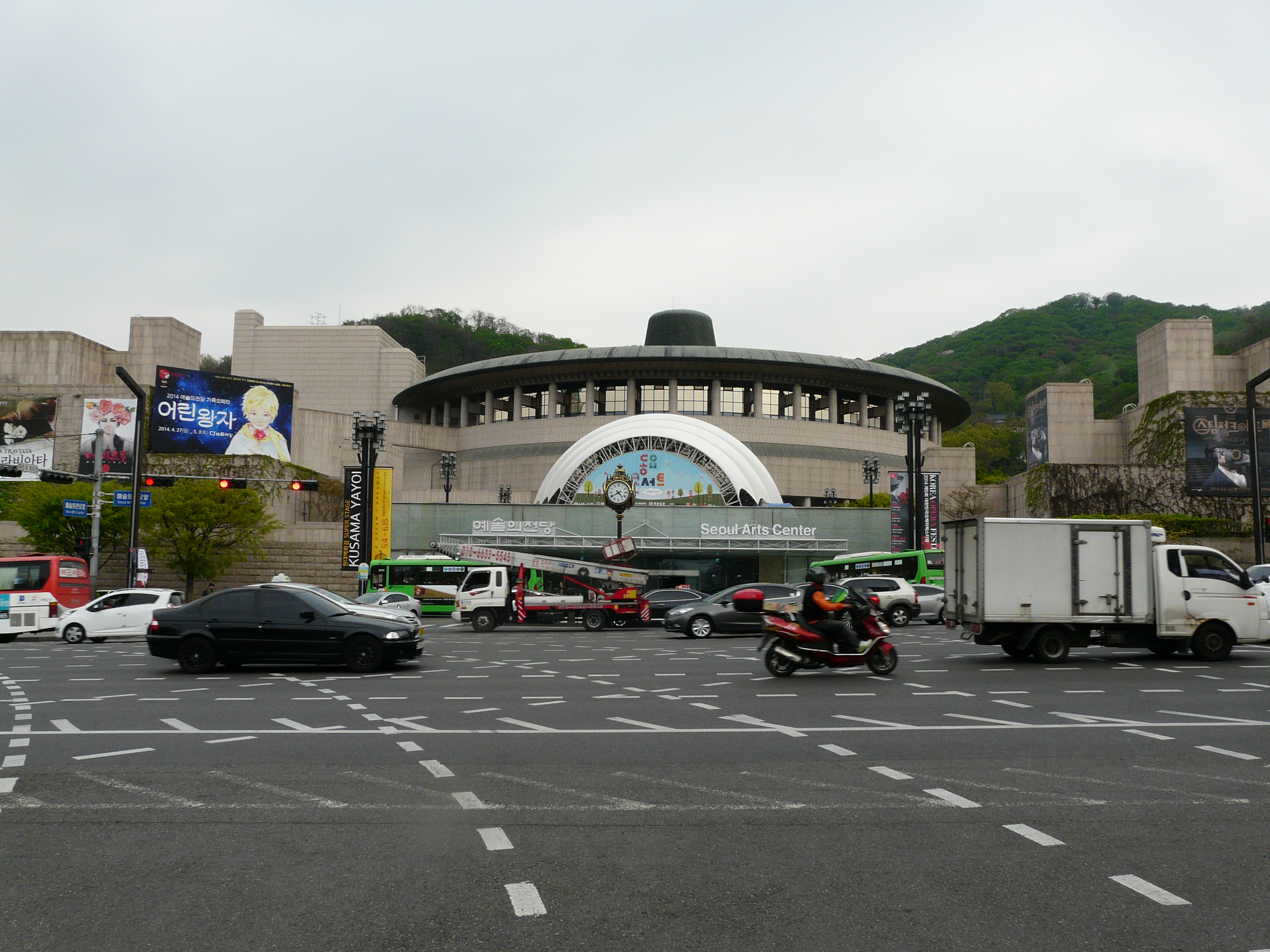 Belebte Kreuzung vor dem Seoul Arts Center in Südkorea, mit vorbeifahrenden Autos, Bussen und Motorrädern und grünen Hügeln und Bannern im Hintergrund.