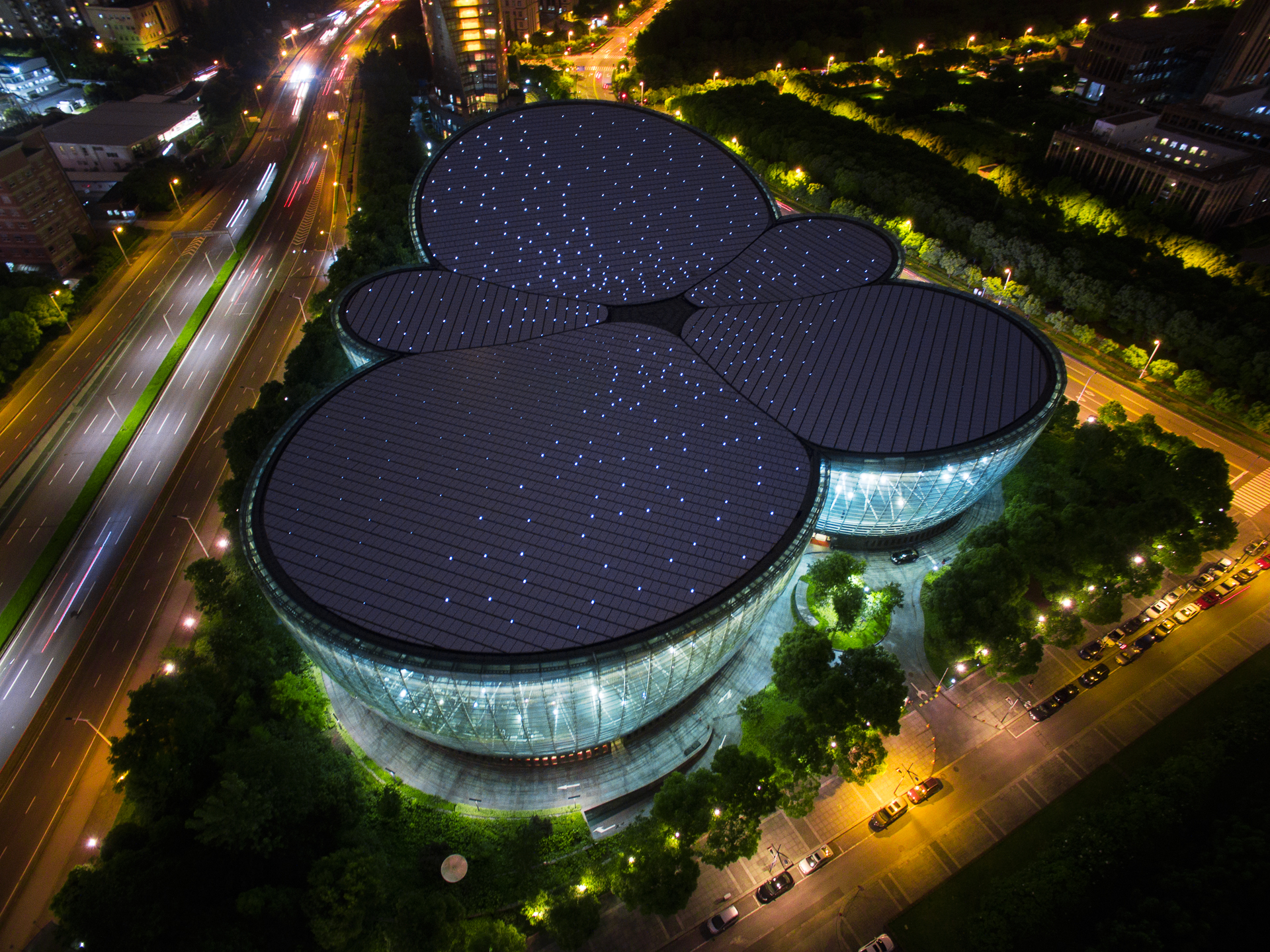 Luftaufnahme eines modernen, einzigartig geformten Gebäudes mit weißem Dach, umgeben von Bäumen und Straßen, in einem städtischen Gebiet mit anderen Gebäuden und Grünflächen in der Nähe.