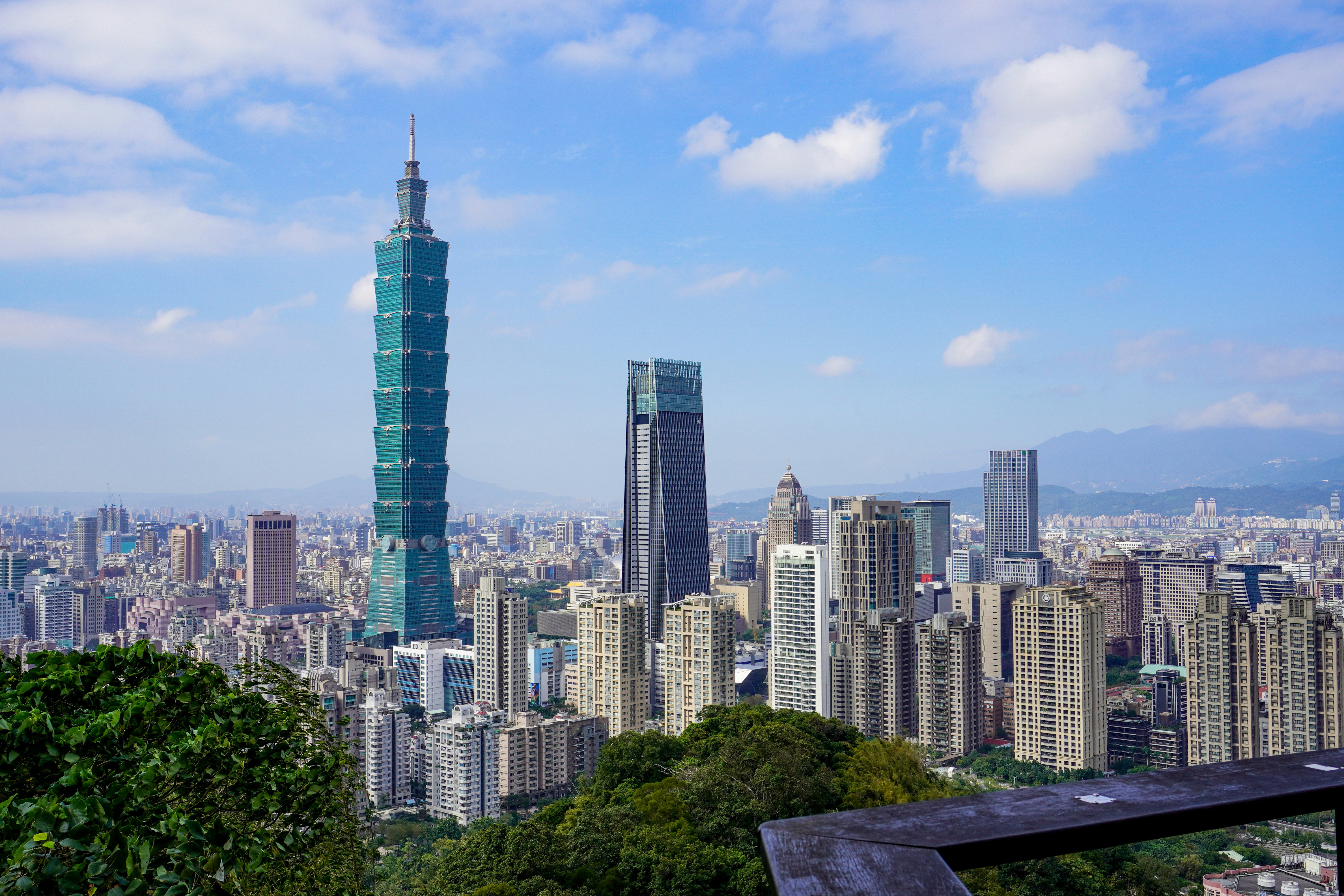 Die Skyline von Taipeh mit dem hohen Wolkenkratzer Taipeh 101, modernen Gebäuden und üppig grünen Bäumen im Vordergrund unter einem blauen Himmel mit vereinzelten Wolken.