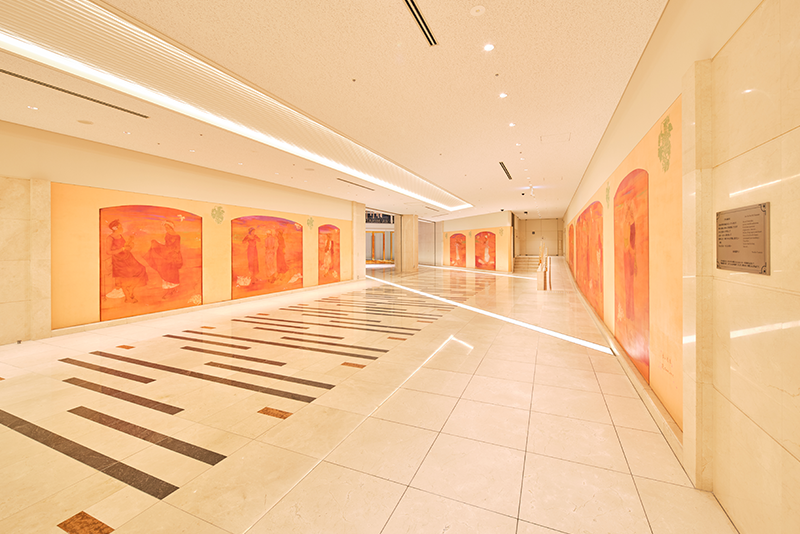 Ein heller, moderner Flur mit hellen Fliesen, abstrakten orangefarbenen Wandbildern an beiden Wänden und in den Boden eingelassenen Linien aus dunklen Fliesen, die zu Glastüren am anderen Ende führen.