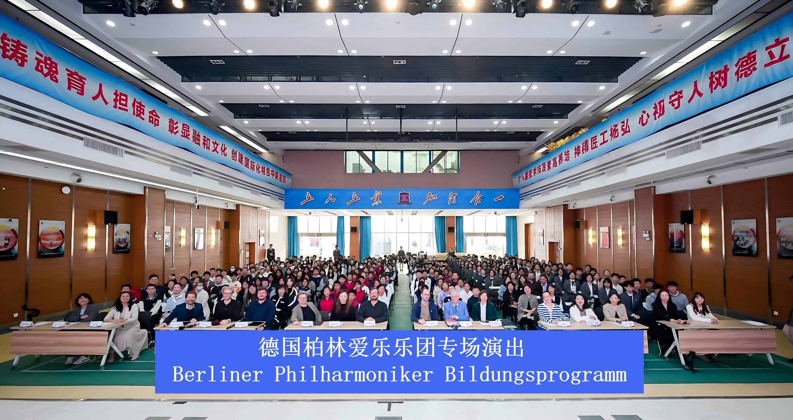 Ein großes Publikum sitzt in einem geräumigen Saal mit Blick auf die Kamera, mit Bannern in chinesischer Sprache und einem blauen Schild mit der Aufschrift "Berliner Philharmoniker Bildungsprogramm" im Vordergrund.