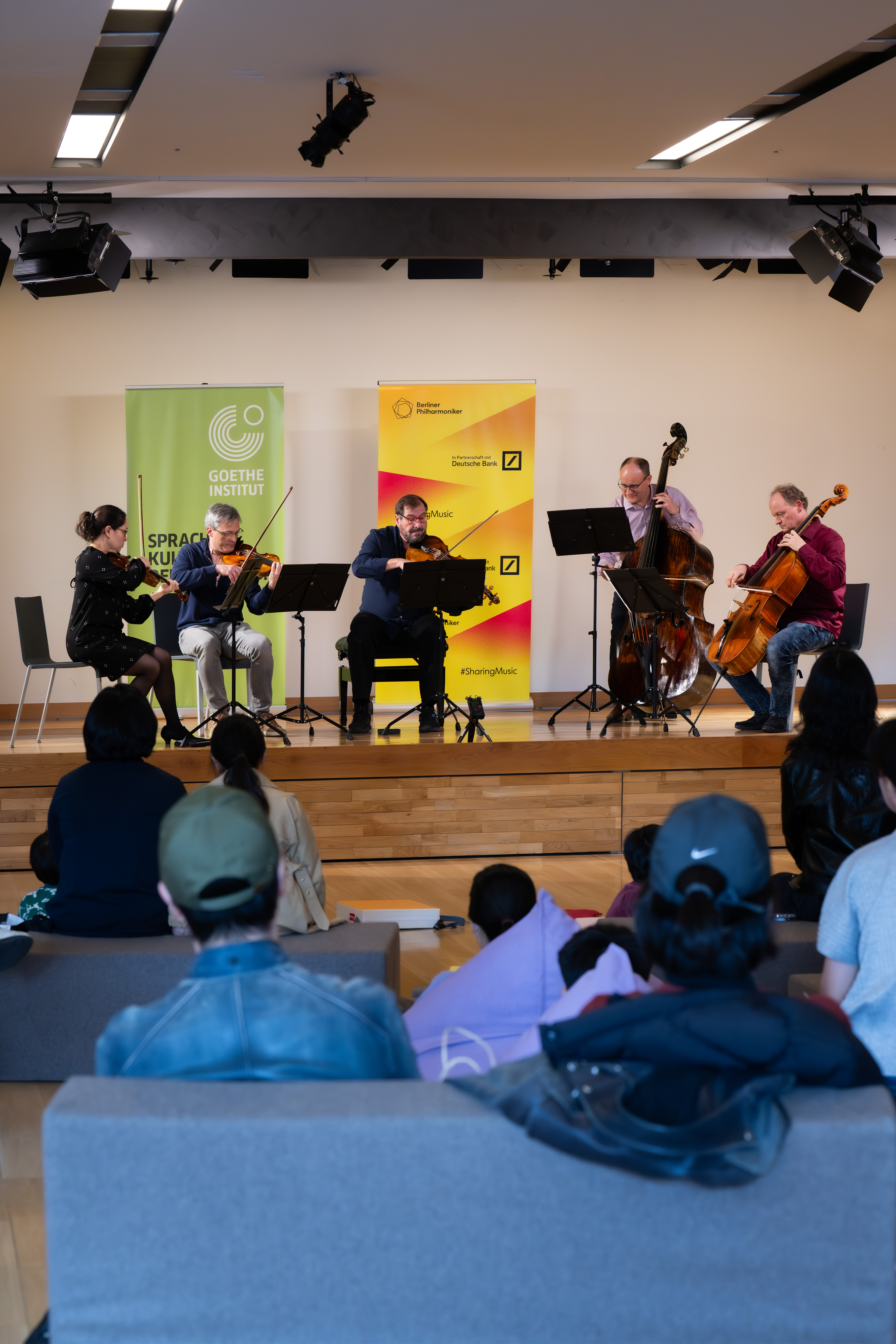 Ein Streichquintett mit Kontrabass tritt auf der Bühne vor einem Publikum auf, hinter dem grüne und gelbe Banner des Goethe-Instituts wehen. Die Zuhörer sitzen auf Sofas und schauen aufmerksam zu.