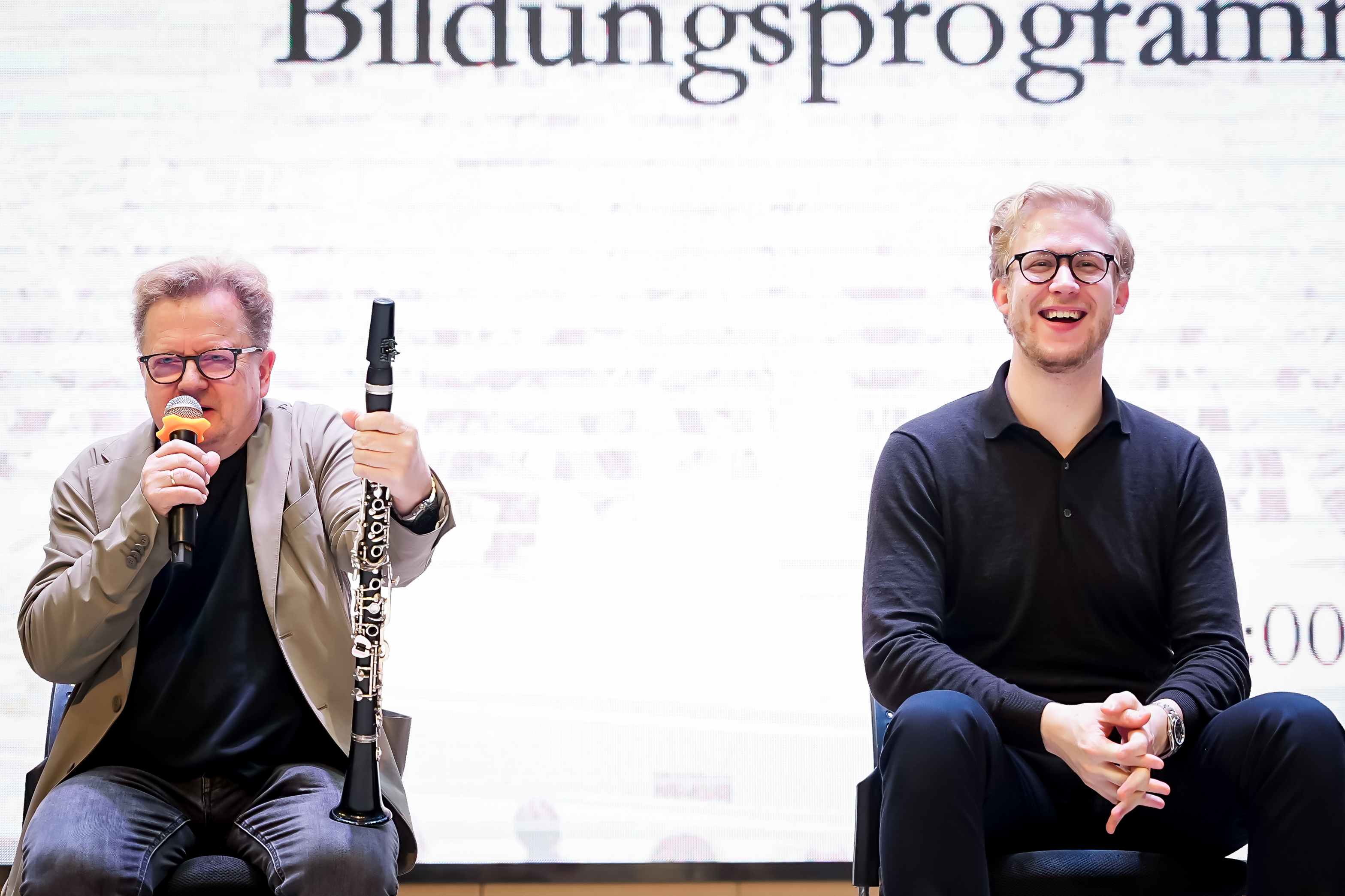 Wenzel Fuchs und Andraž Golob sitzen auf Stühlen vor einem hellen Bildschirm. Wenzel Fuchs, der eine Klarinette und ein Mikrofon in der Hand hält, spricht. Andraž Golob, der eine Brille trägt, sitzt lächelnd und lachend da.