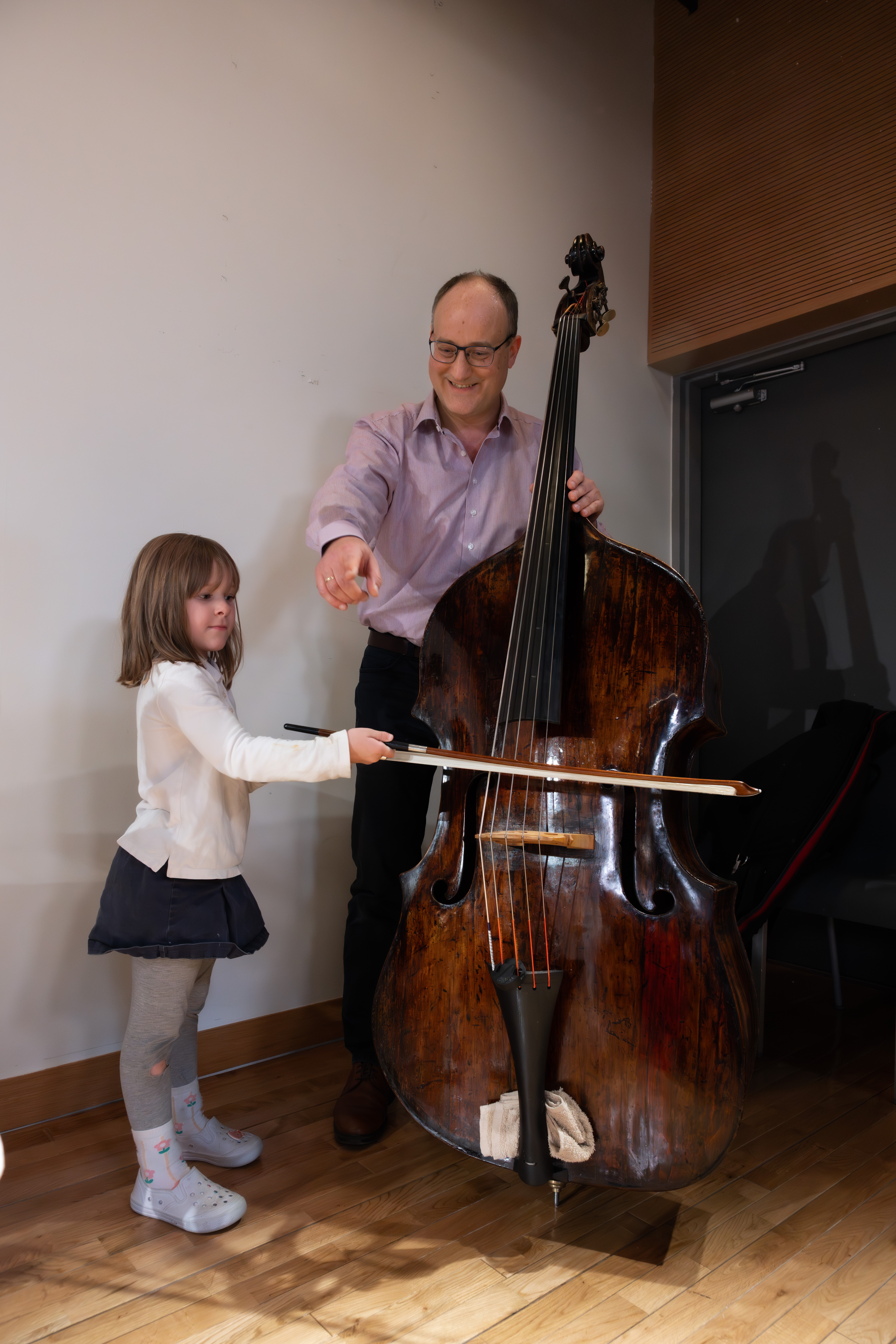 Ein lächelnder Janusz Widzyk hilft einem jungen Mädchen in einem hellen Raum mit Holzboden, einen Kontrabass mit einem Bogen zu spielen. Das Mädchen schaut konzentriert, während der Mann ihre Hand auf dem Instrument führt.