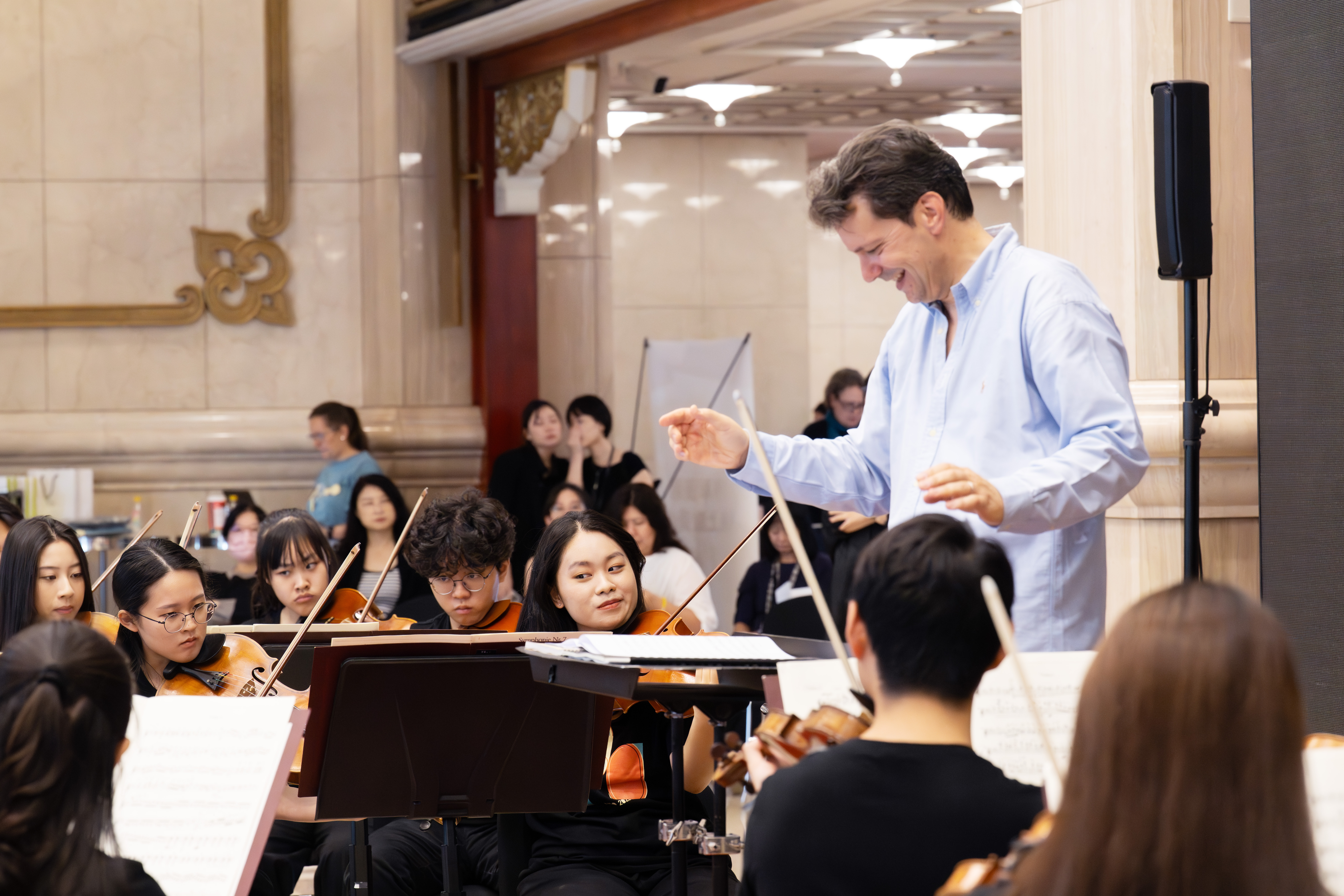 Geiger Simon Bernardini im hellblauen Hemd leitet ein Orchester aus jungen Musikern, die in einer prächtig geschmückten Halle Streichinstrumente spielen.