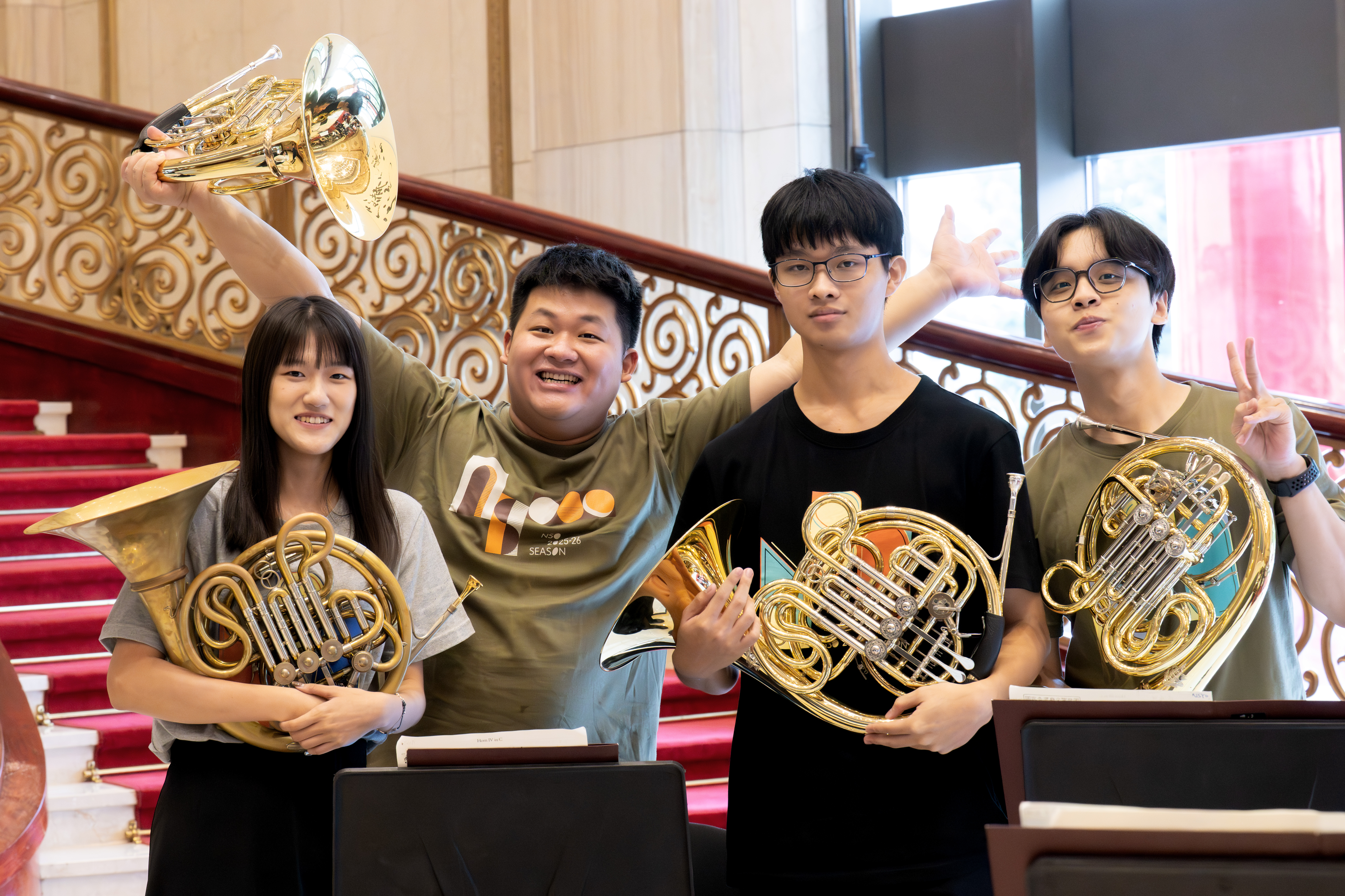 Solohornist Yun Zeng und drei junge Hornisten lächeln und posieren vor einer mit rotem Teppich ausgelegten Treppe mit kunstvollem Geländer. 
