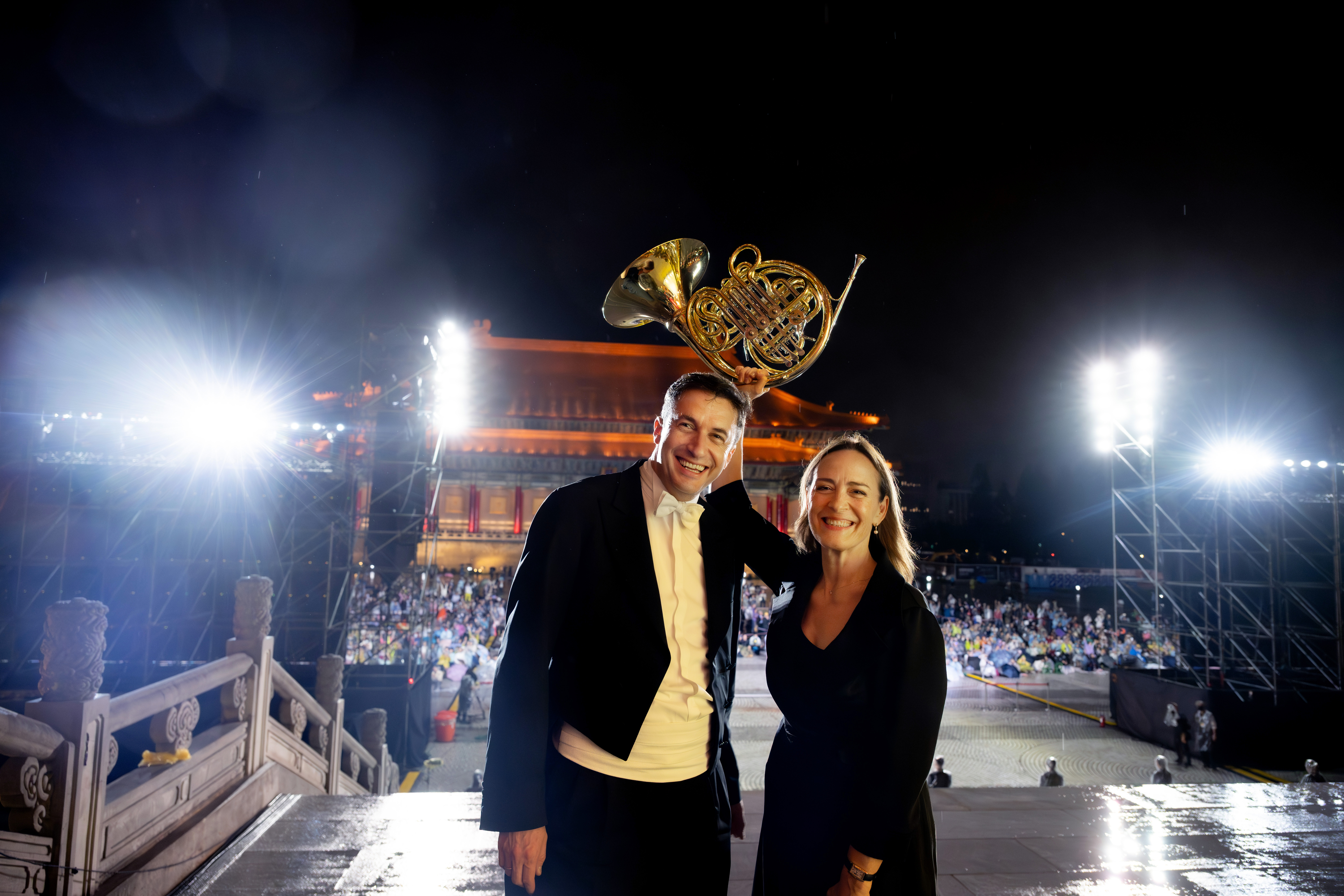 Noah Bendix-Balgley und Sarah Willis draußen beim public viewing in Taipeh, Sarah Willis hält ihr Horn hoch in die Luft, mit hellen Bühnenlichtern und einer großen Menschenmenge vor einem beleuchteten Gebäude bei Nacht.