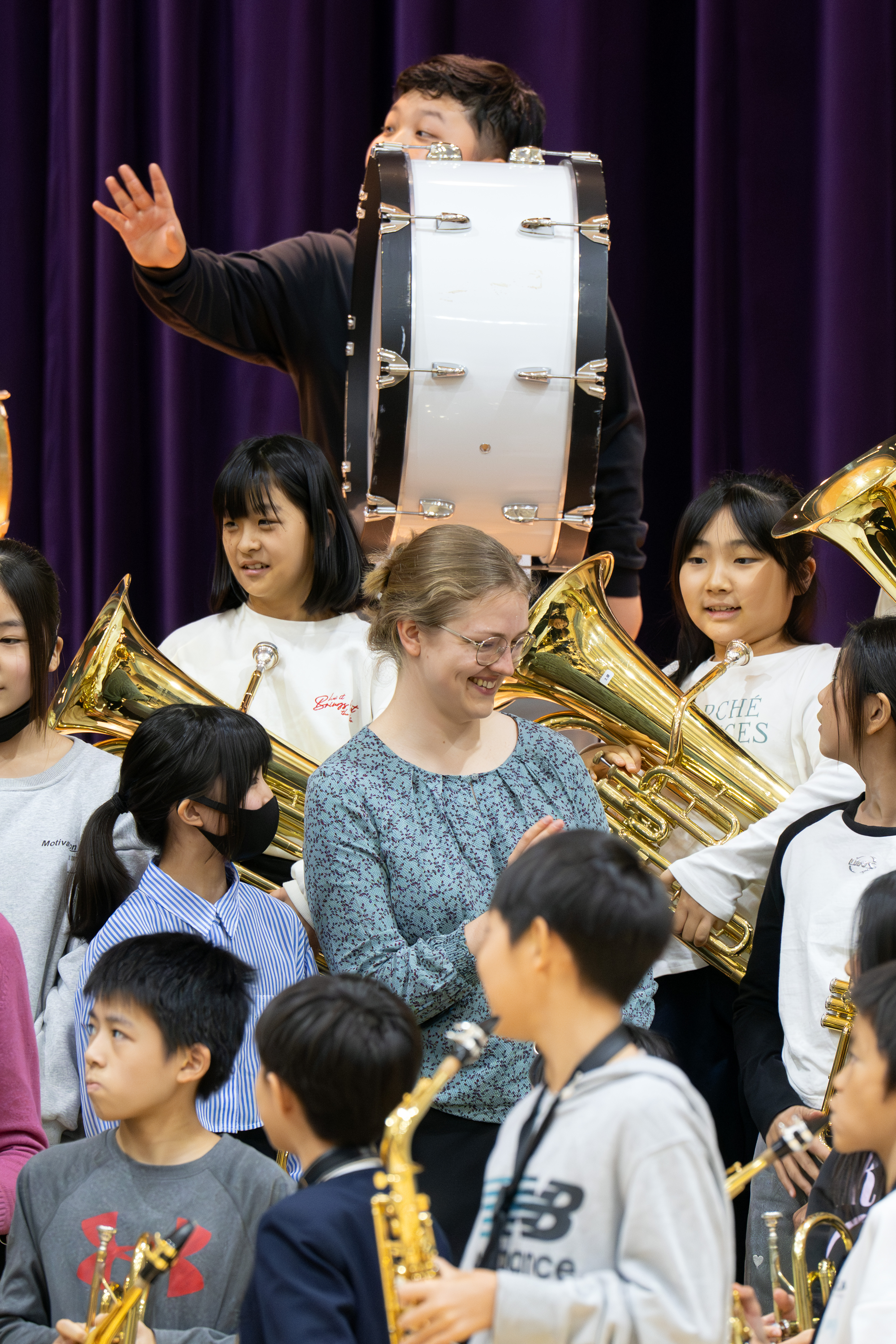 Eine Gruppe von Kindern mit Blasinstrumenten und Hornistin Paula Ernesaks stehen zusammen, lächeln und unterhalten sich. Hinter ihnen spielt eine Person eine große Basstrommel. Im Hintergrund hängt ein lila Vorhang.