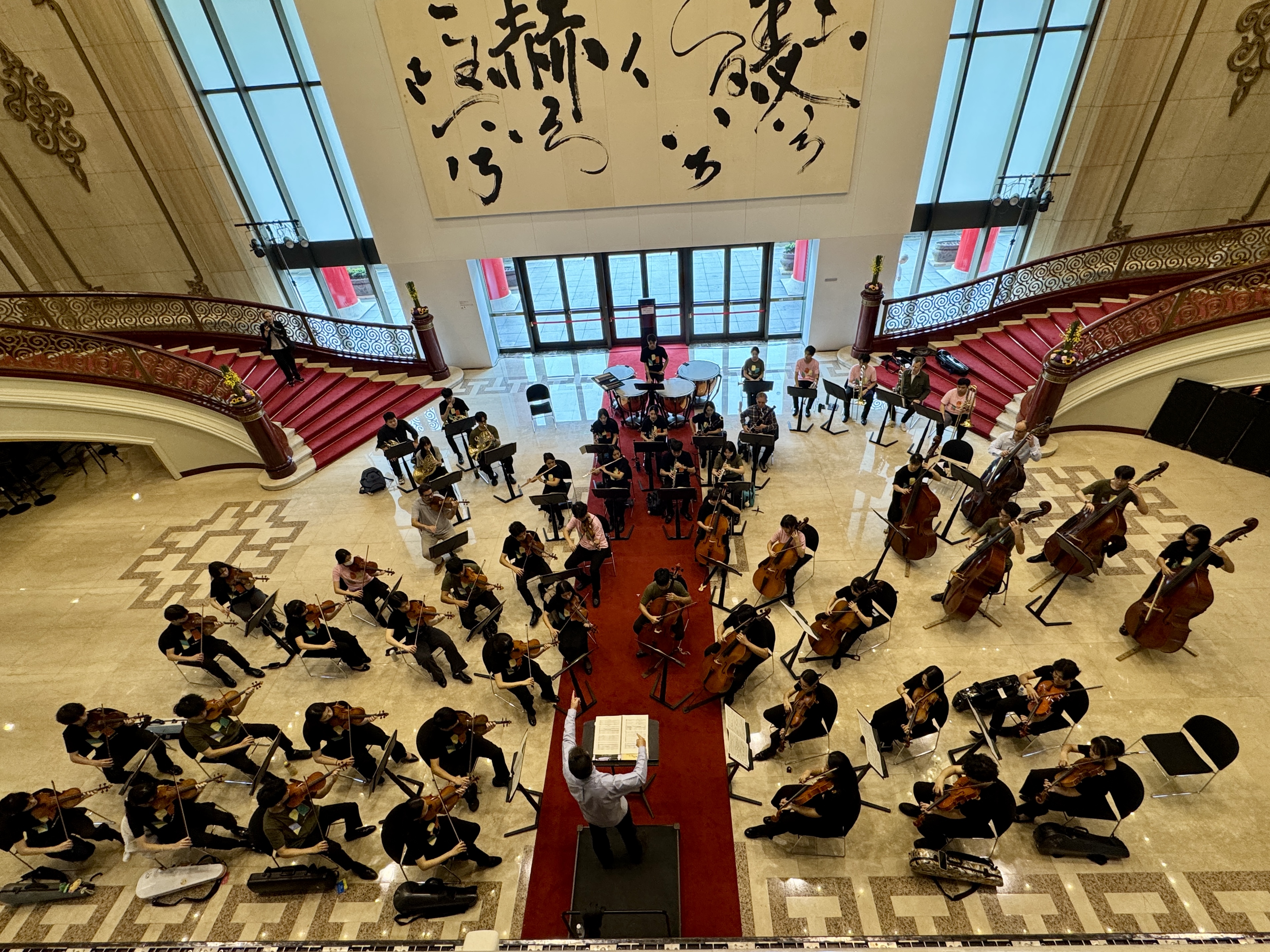 Blick von oben auf ein Orchester, das in einem großen Saal mit rotem Teppich, geschwungenen Treppen und großen Fenstern probt. Ein Dirigent steht in der Mitte, Musiker spielen Streichinstrumente, und darüber hängt ein großes Kunstwerk.