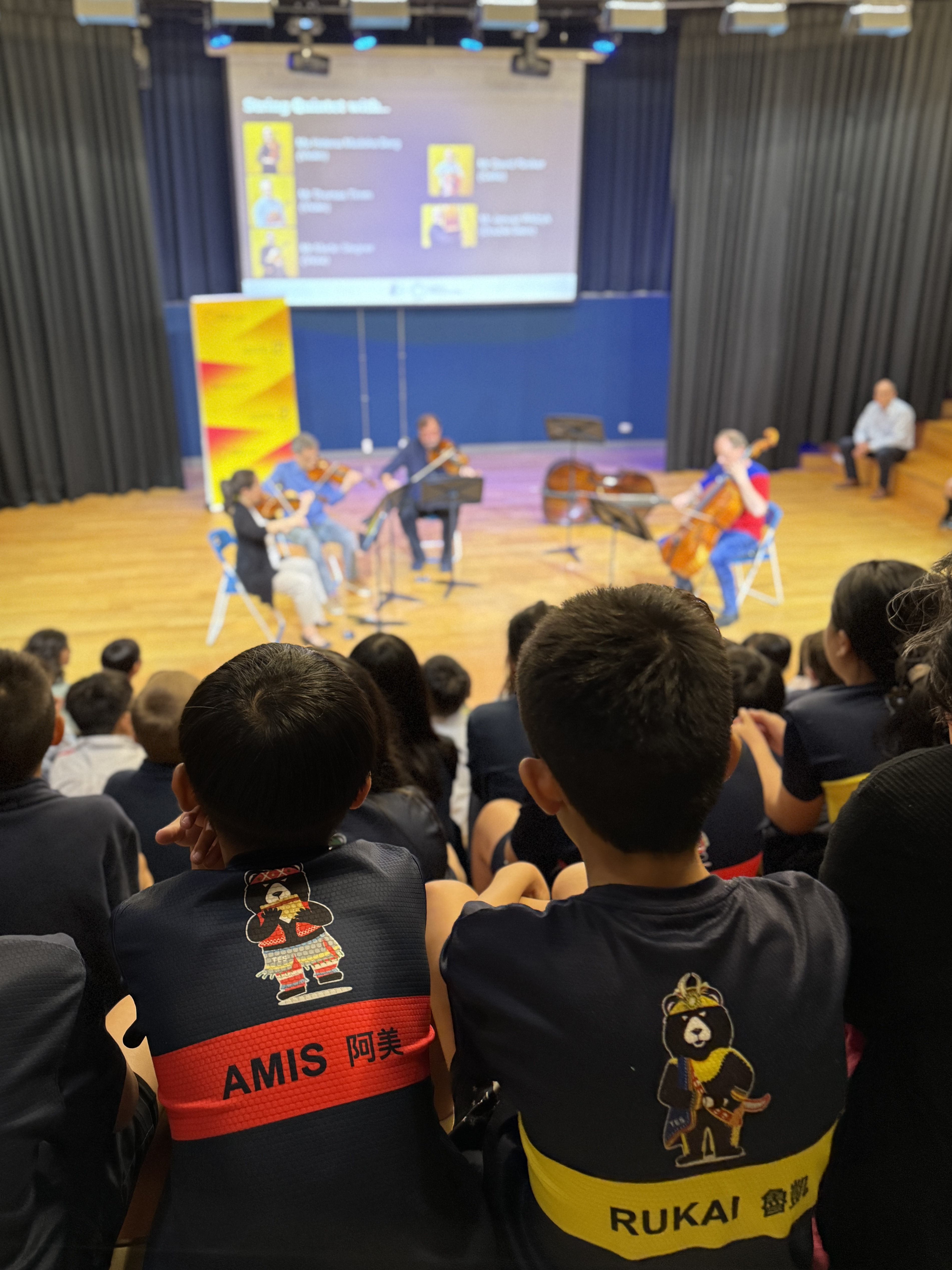 Kinder beobachten den Auftritt eines Streichquartetts auf einer Bühne in einer Schulaula. Zwei Kinder im Vordergrund tragen Hemden mit der Aufschrift AMIS und RUKAI und Cartoon-Figuren auf dem Rücken.