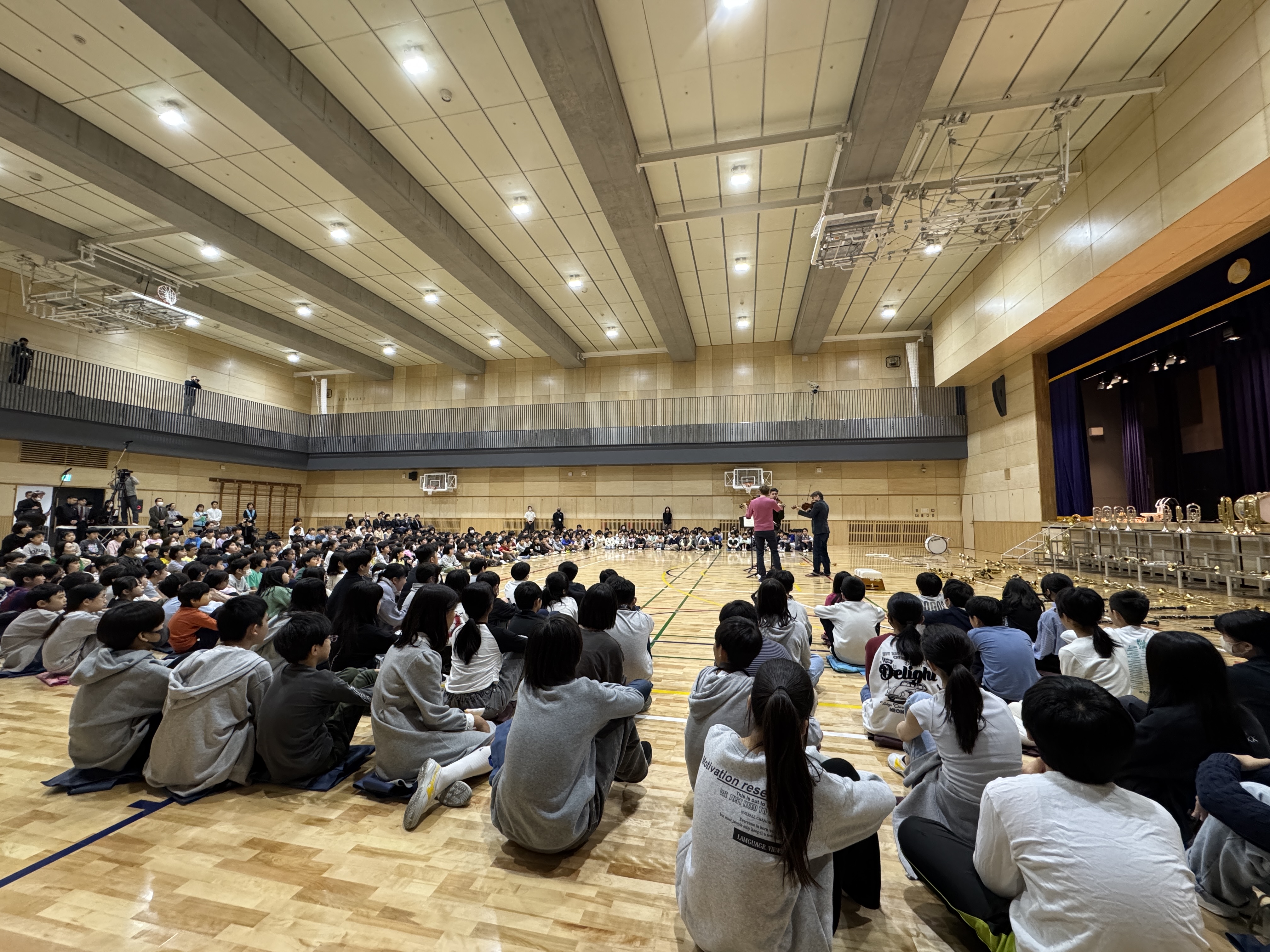 Eine große Gruppe von Schülern sitzt auf dem Boden einer Turnhalle und blickt auf eine Bühne, auf der einige Musiker stehen. Auf der Bühne sind Stuhlreihen und Musikinstrumente unter hellen Scheinwerfern zu sehen.