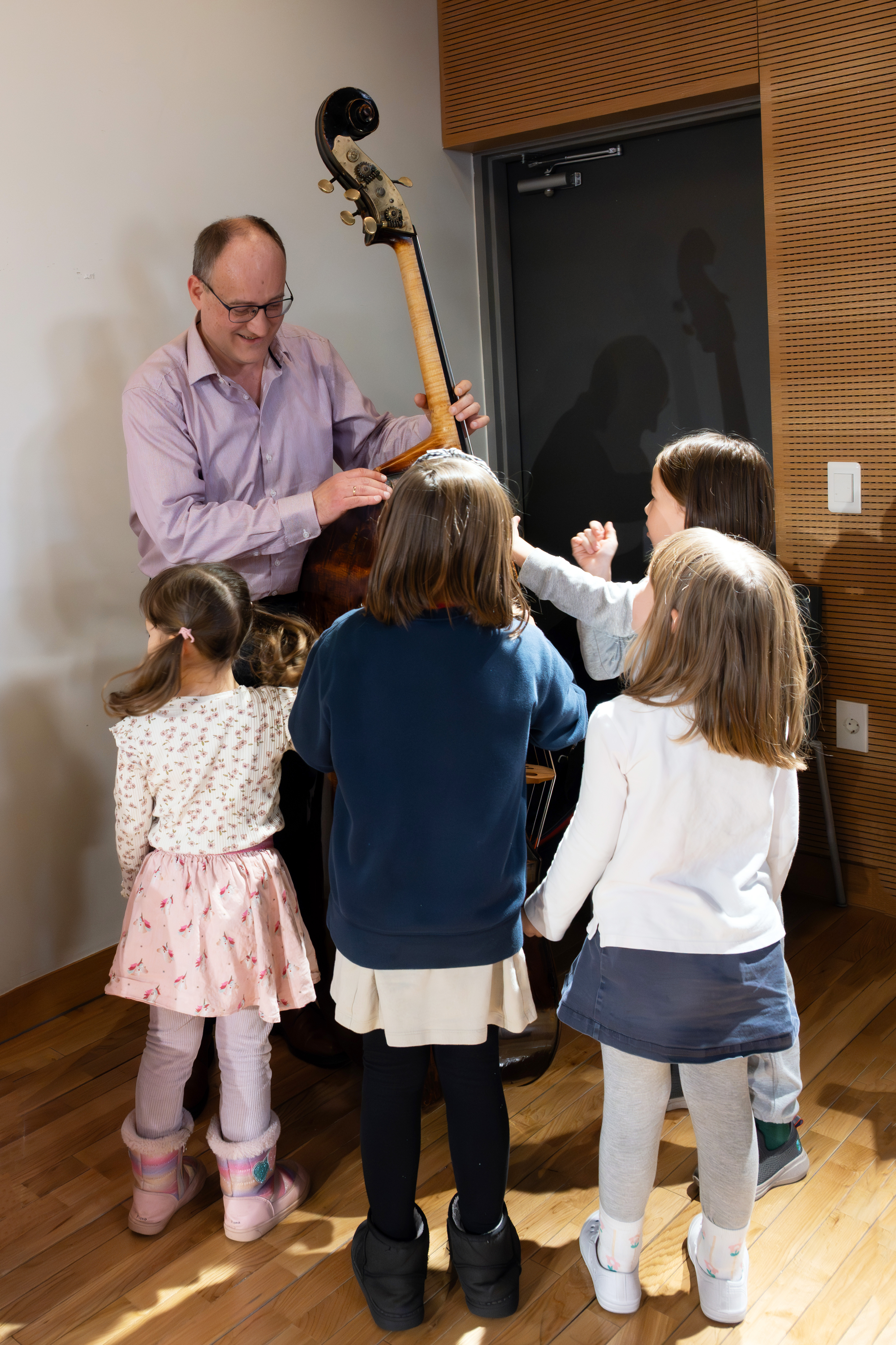 A smiling man demonstrates a double bass to a group of five young girls gathered around him in a well-lit room with wooden floors.