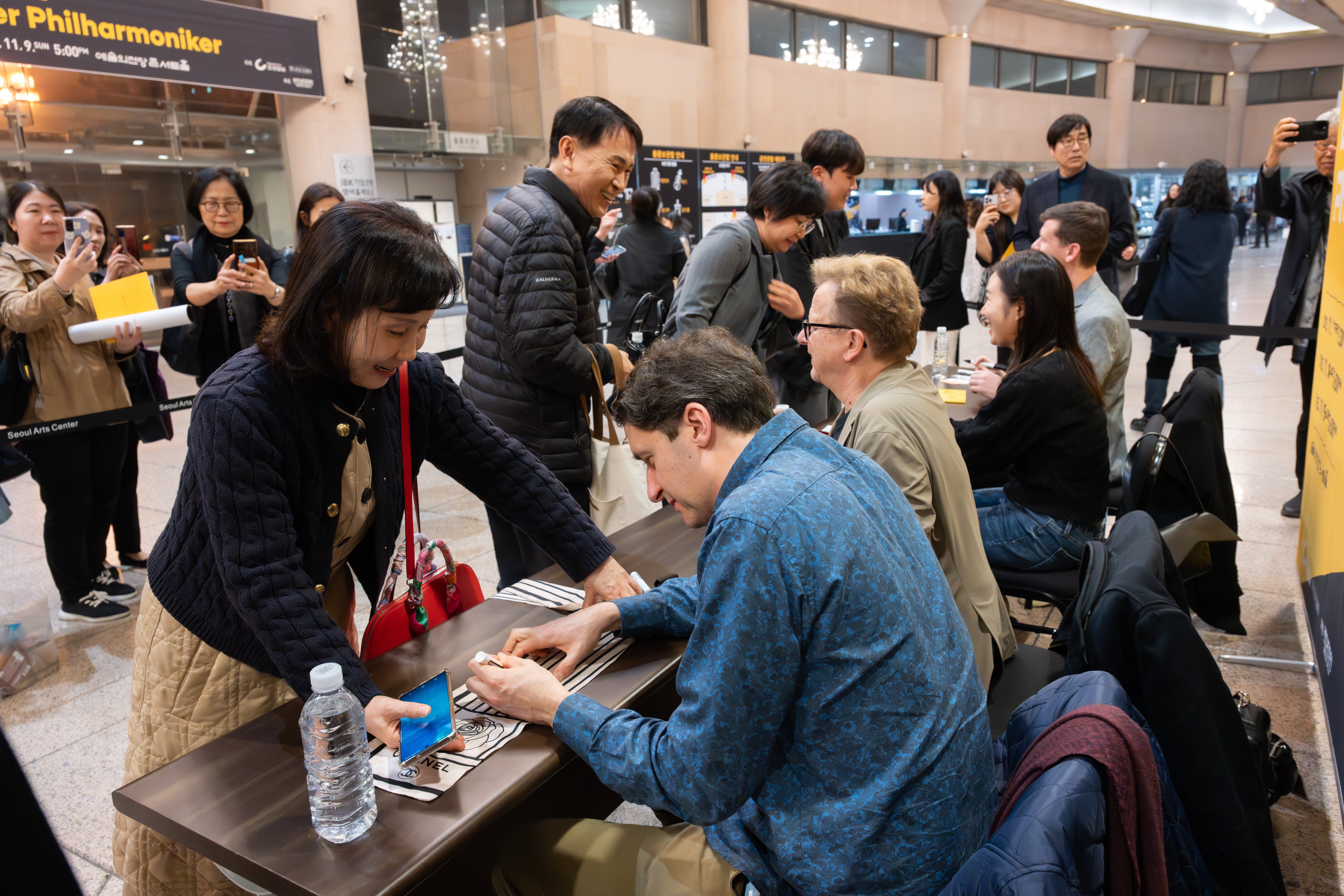 People gather around a table for an autograph session; musicians sign programs for fans, while others wait in line and take photos in a busy indoor venue.