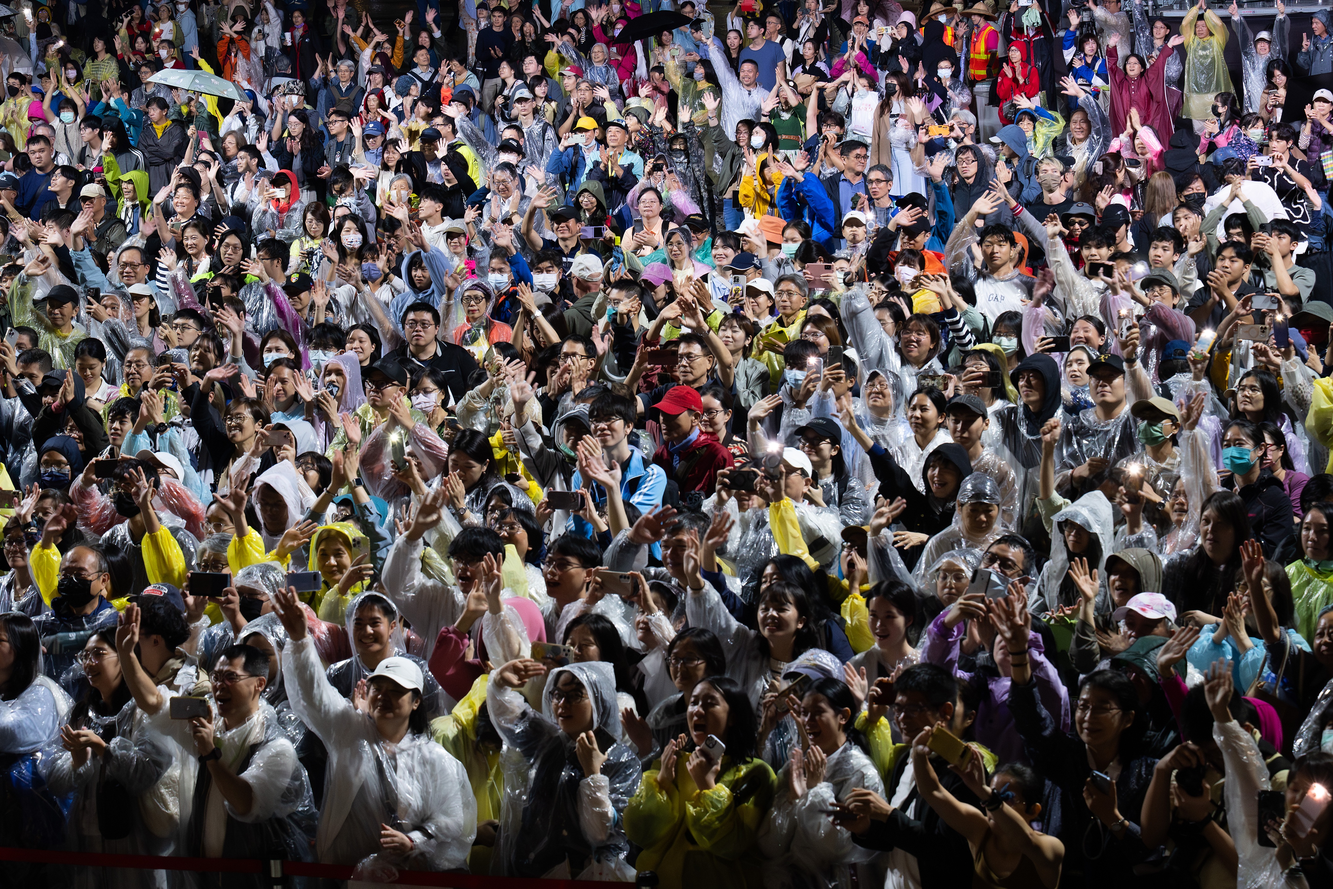 A large crowd of people, many wearing rain ponchos and hats, cheer and clap enthusiastically at an outdoor event, despite the wet weather.