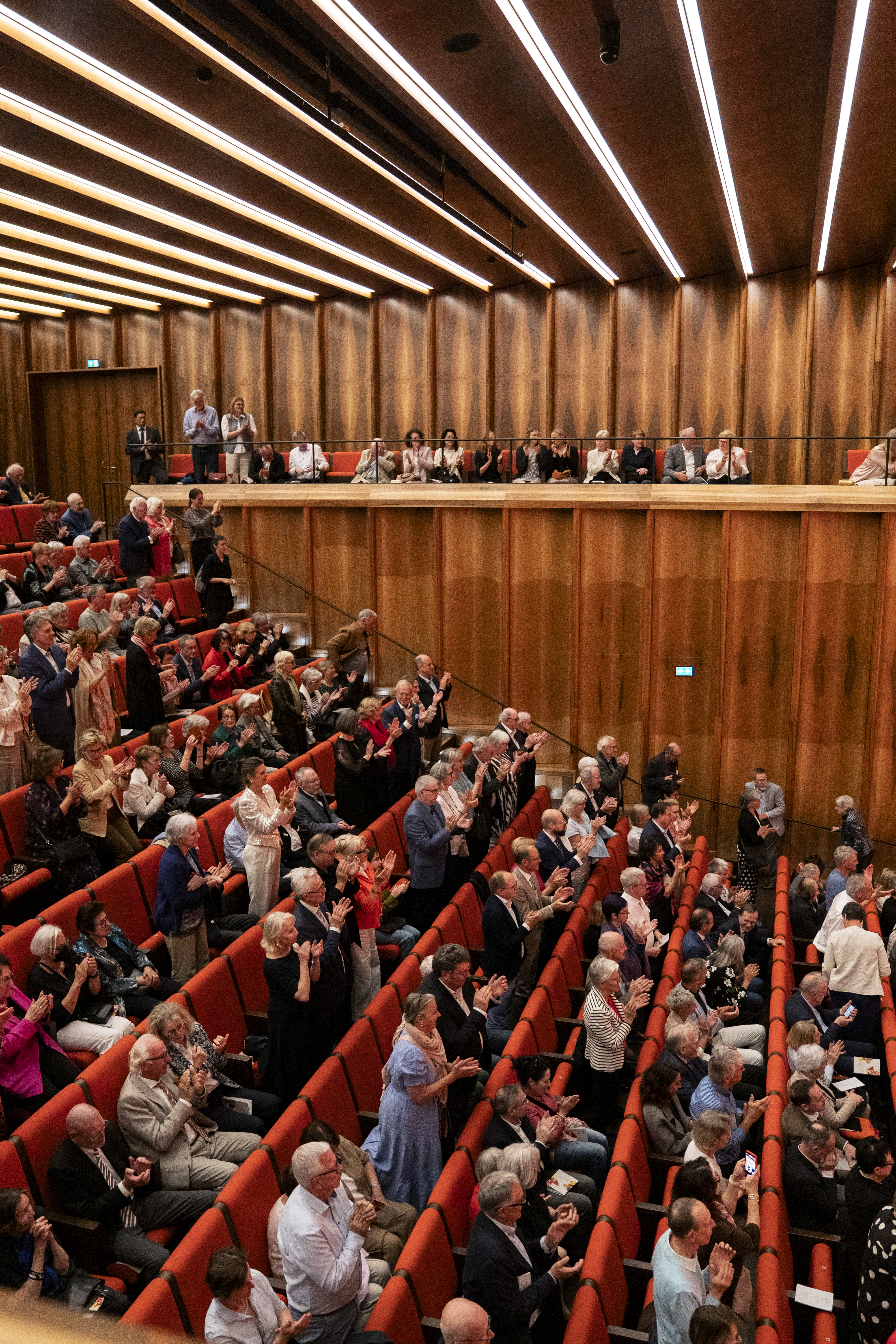 Blick in die voll besetzten Zuschauerreihen eines modernen Konzertsaals. Das Publikum sitzt in roten Sesseln, steht teilweise und applaudiert begeistert. Die Seitenwände und die Decke sind mit hellem Holz gestaltet, was dem Raum eine edle und warme Atmosphäre verleiht.