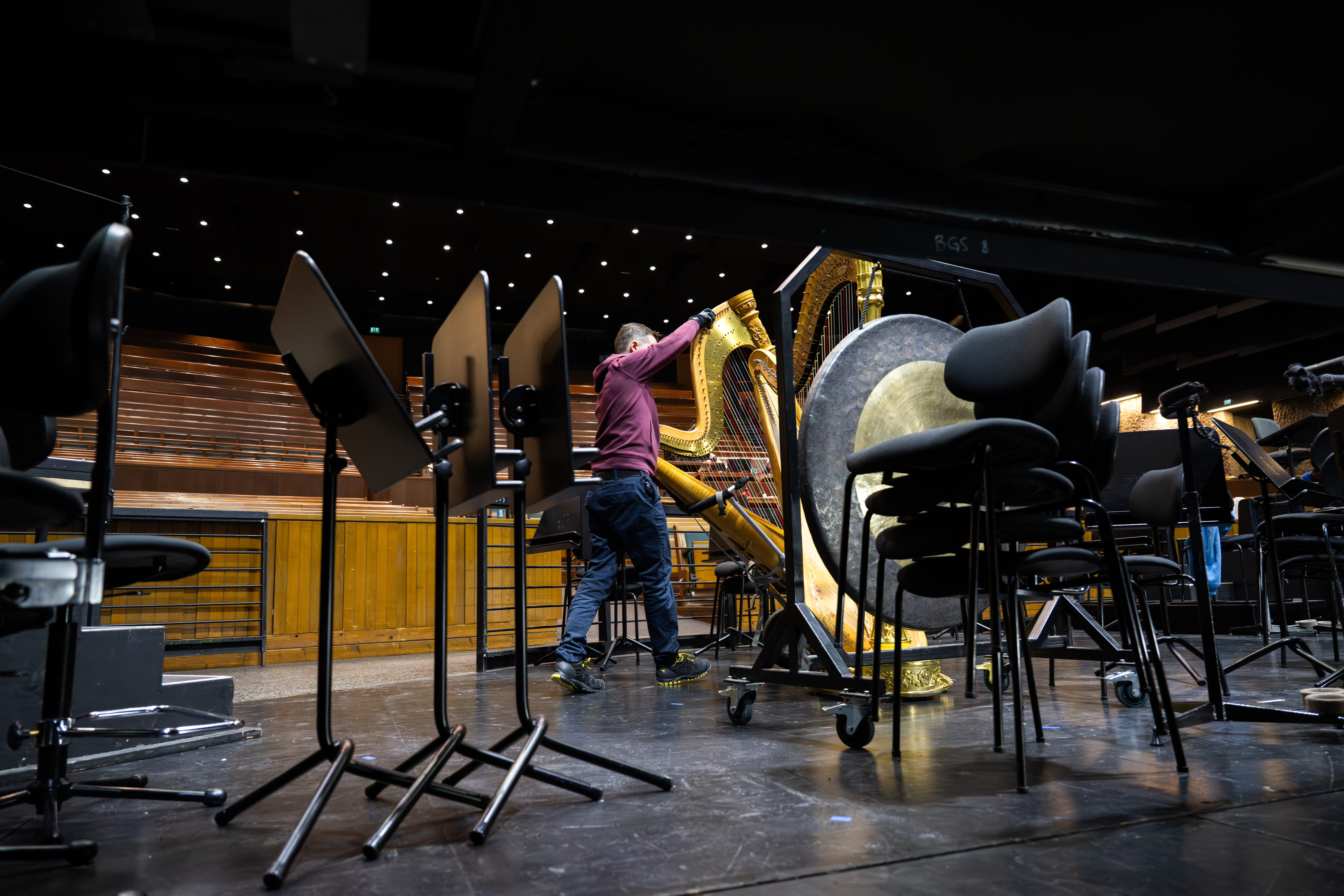 Eine Person justiert eine Harfe auf der Bühne eines Konzertsaals, umgeben von leeren Notenständern und gestapelten Stühlen, um sich auf einen Auftritt vorzubereiten.