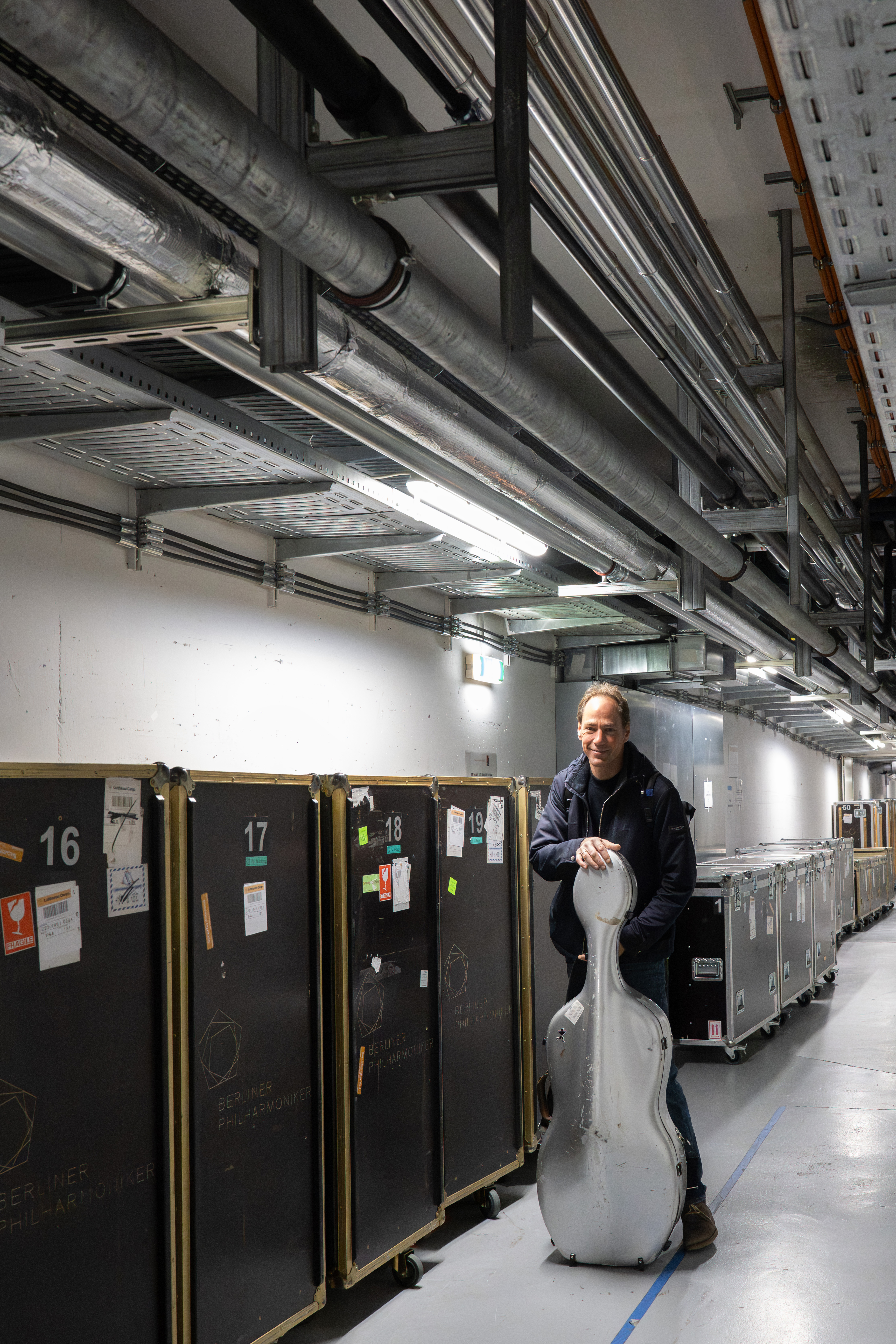 Ein Mann steht in einem Industrieflur neben großen schwarzen Lagerkästen und hält einen silbernen Cellokasten aufrecht.