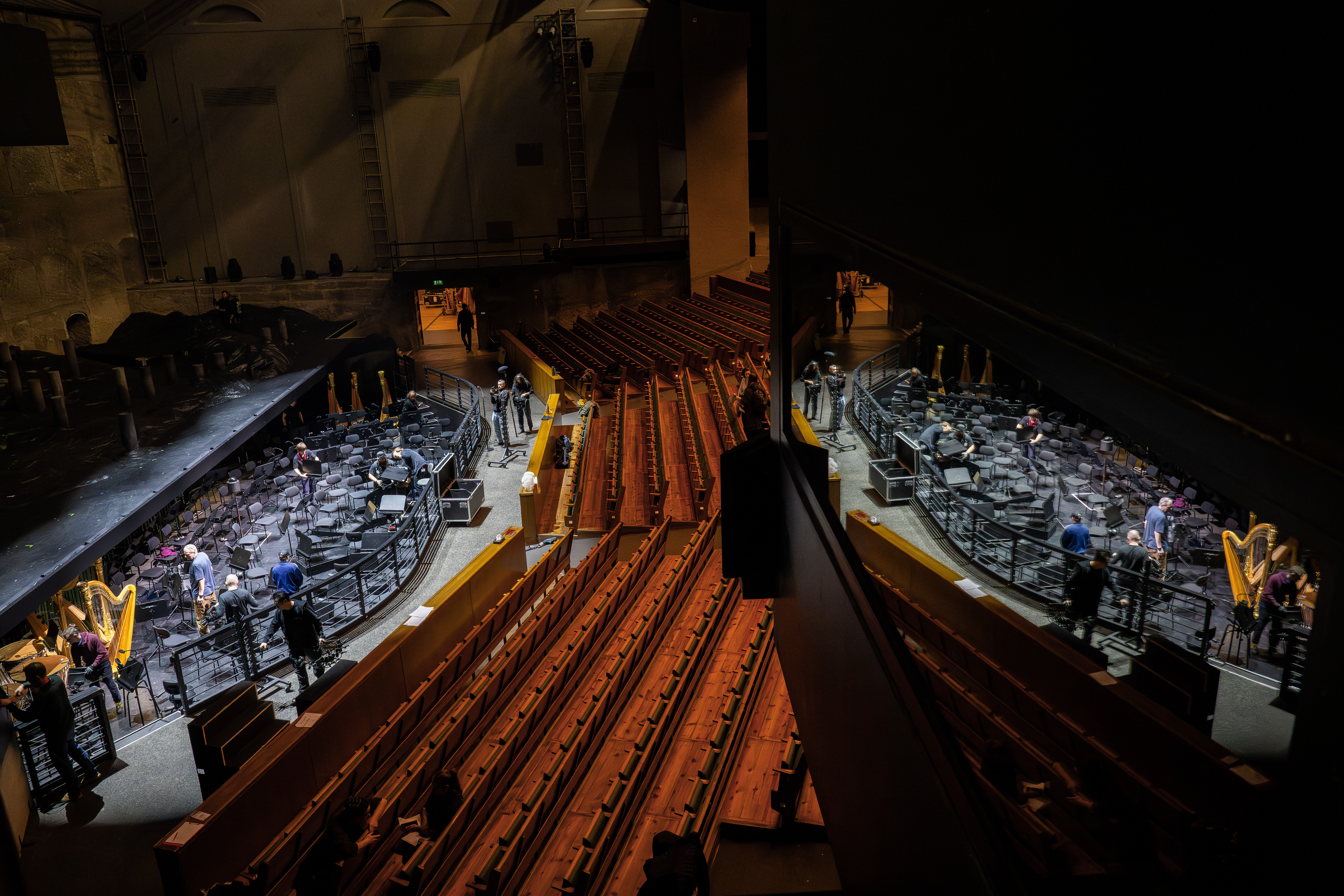 Blick auf einen leeren Theatersaal mit Holzsitzen und einem Orchestergraben, in dem Musiker und Harfen zu sehen sind. Die Szene wird in einem nahe gelegenen Spiegel reflektiert, was den visuellen Effekt verdoppelt.