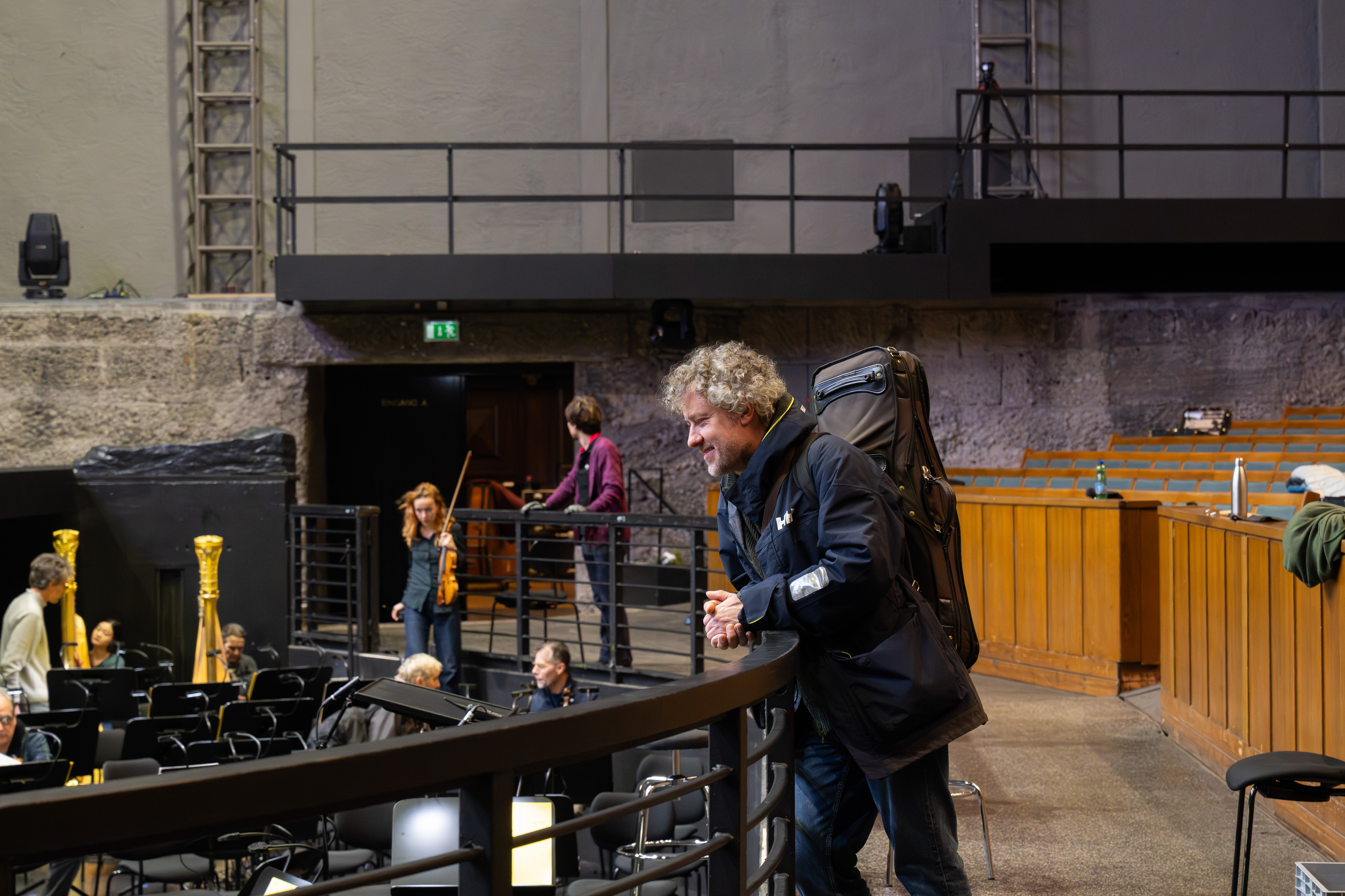Ein Mann mit lockigem Haar und einem Geigenkasten auf dem Rücken lehnt lächelnd an einem Geländer im Theater und blickt in den Orchestergraben, während sich im Hintergrund Musiker und andere Personen an ihren Instrumenten vorbereiten.