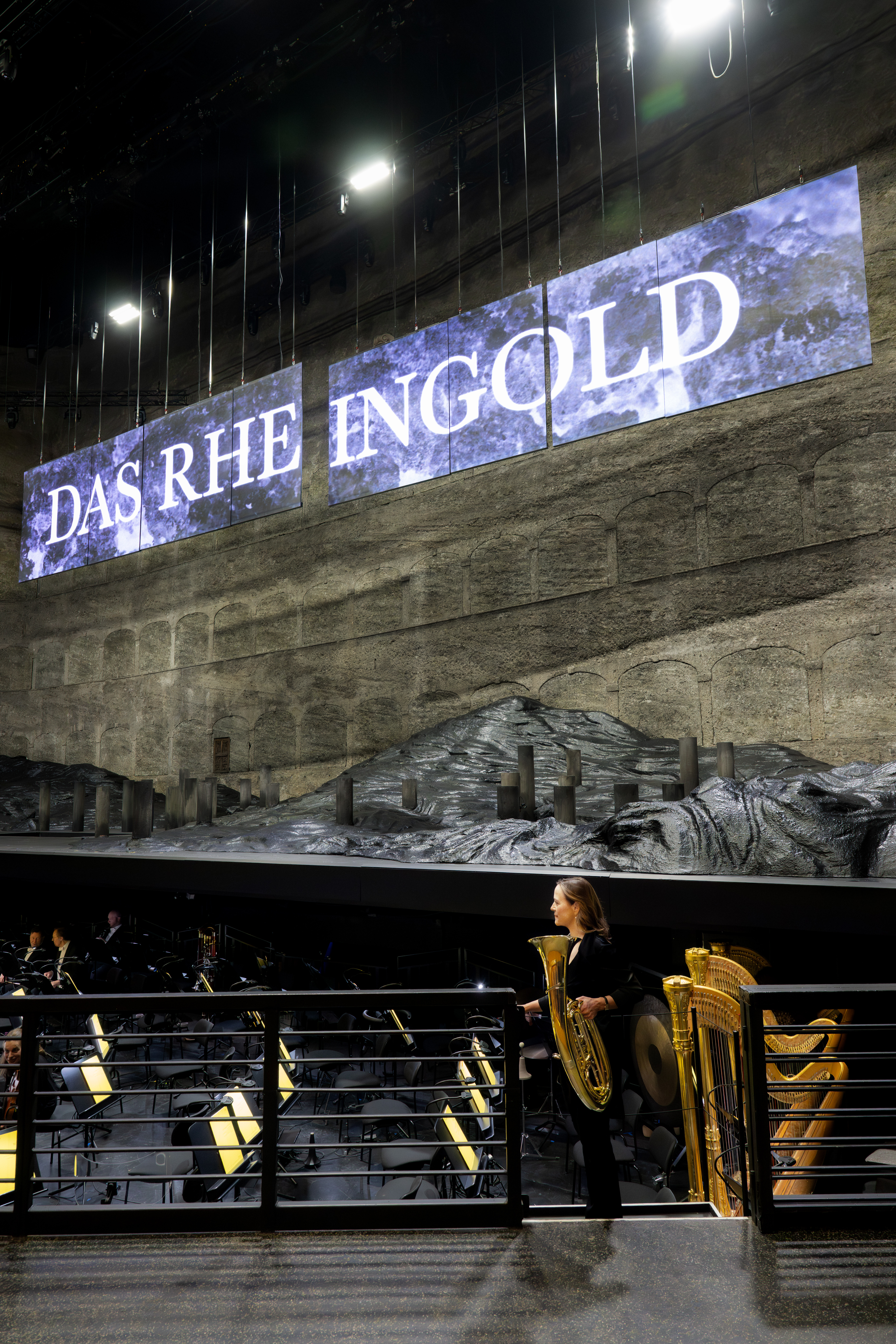 Hornistin Sarah Willis betritt den Orchestergraben vor der Vorstellung, im Hintergrund sieht man die Bühne, sowie Bildschirme mit der Aufschrift »Das Rheingold«.