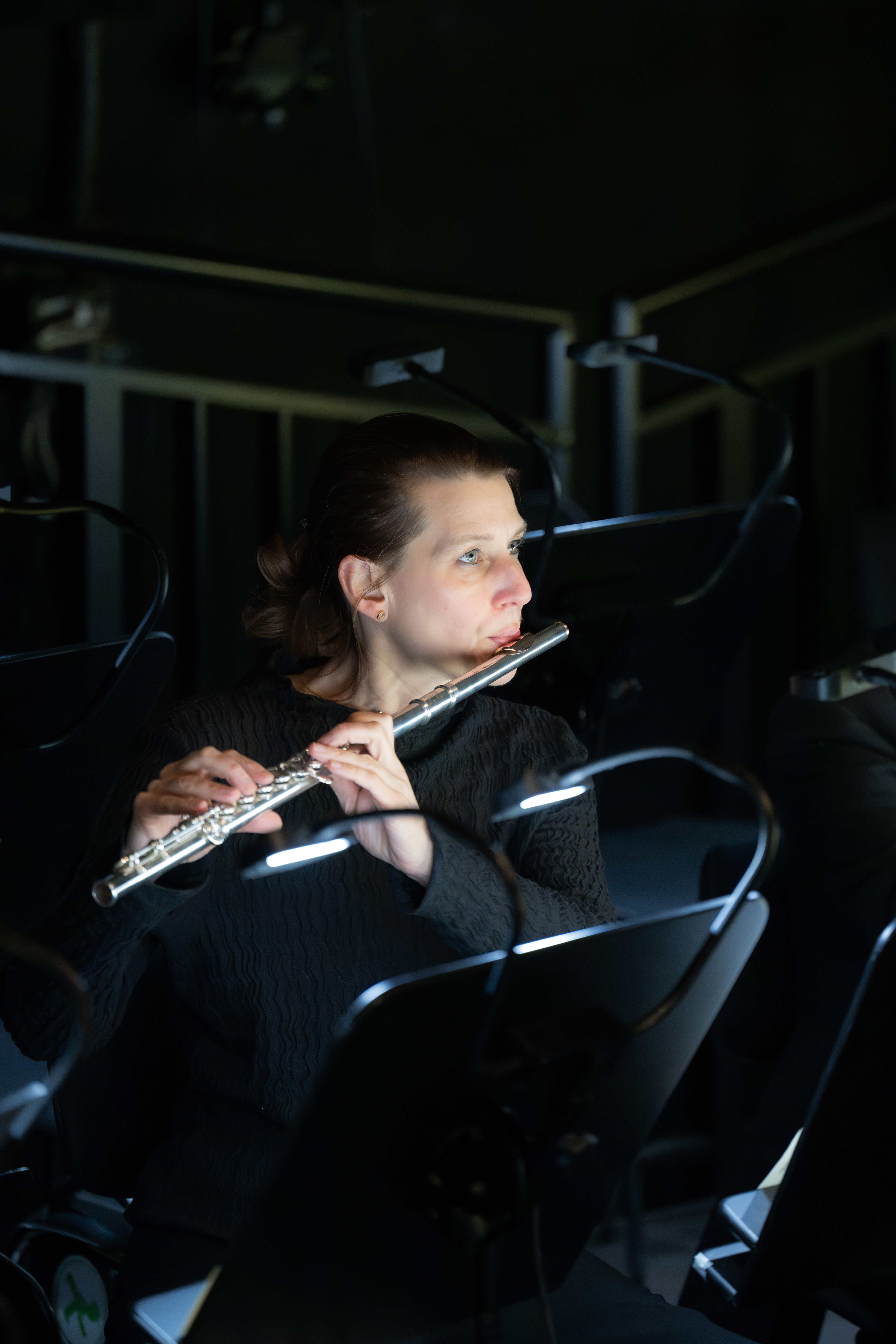 Flötistin Jelka Weber, in schwarz gekleidet mit kurzen Haaren, spielt Flöte in einem schwach beleuchteten Orchestergraben, umgeben von Notenständern mit kleinen Lichtern.