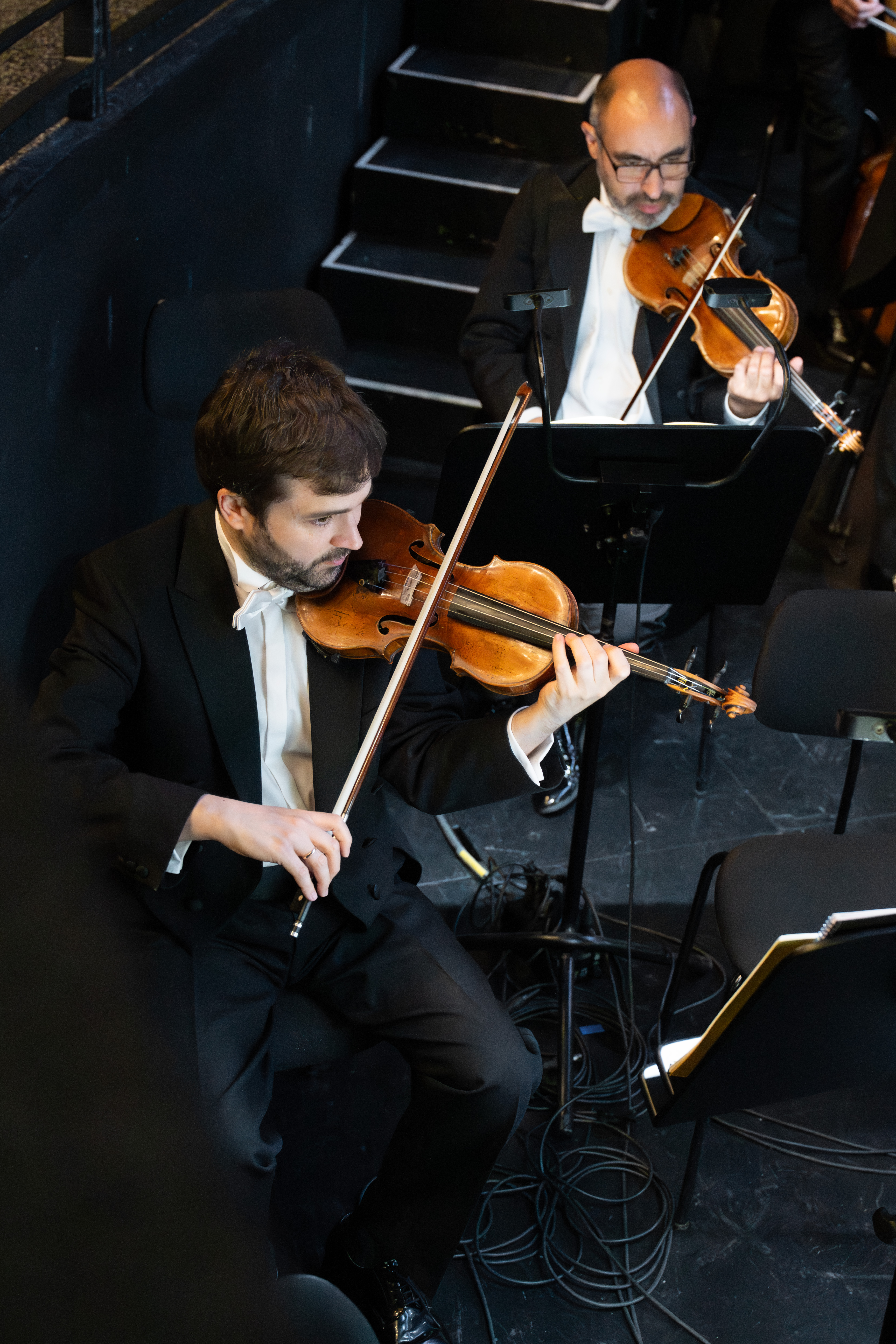 Bratscher Allan Nilles und Geiger Álvaro Parra in formellen schwarzen Anzügen und weißen Fliegen spielen in einem Orchestergraben, mit Notenständern vor ihnen und schwarzen Treppen im Hintergrund.