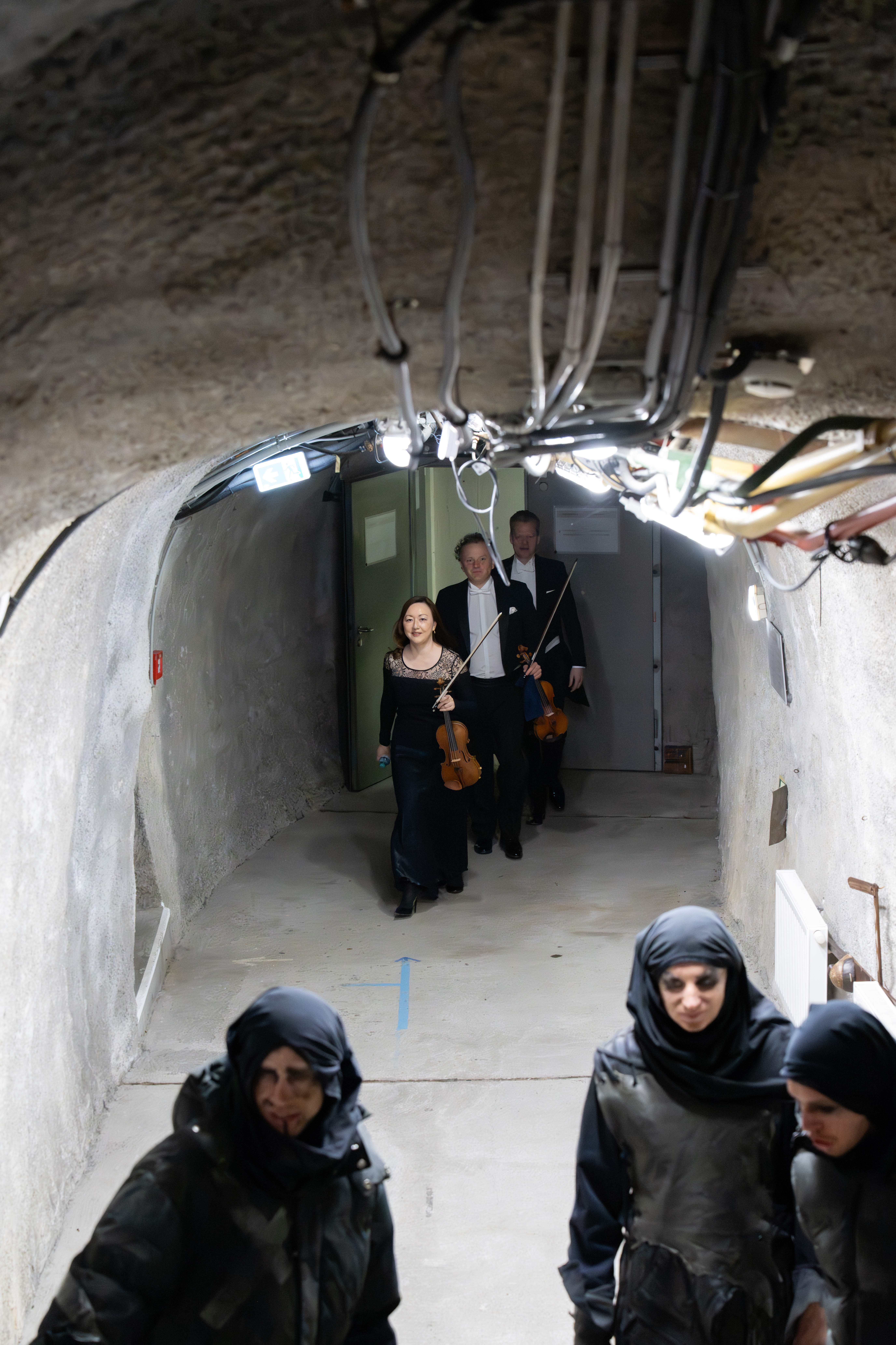 Musiker in förmlicher Kleidung mit Geigen in der Hand gehen durch einen unterirdischen Tunnel, während im Vordergrund Menschen in dunkler taktischer Kleidung mit Kapuzen und Gesichtsbemalung stehen.