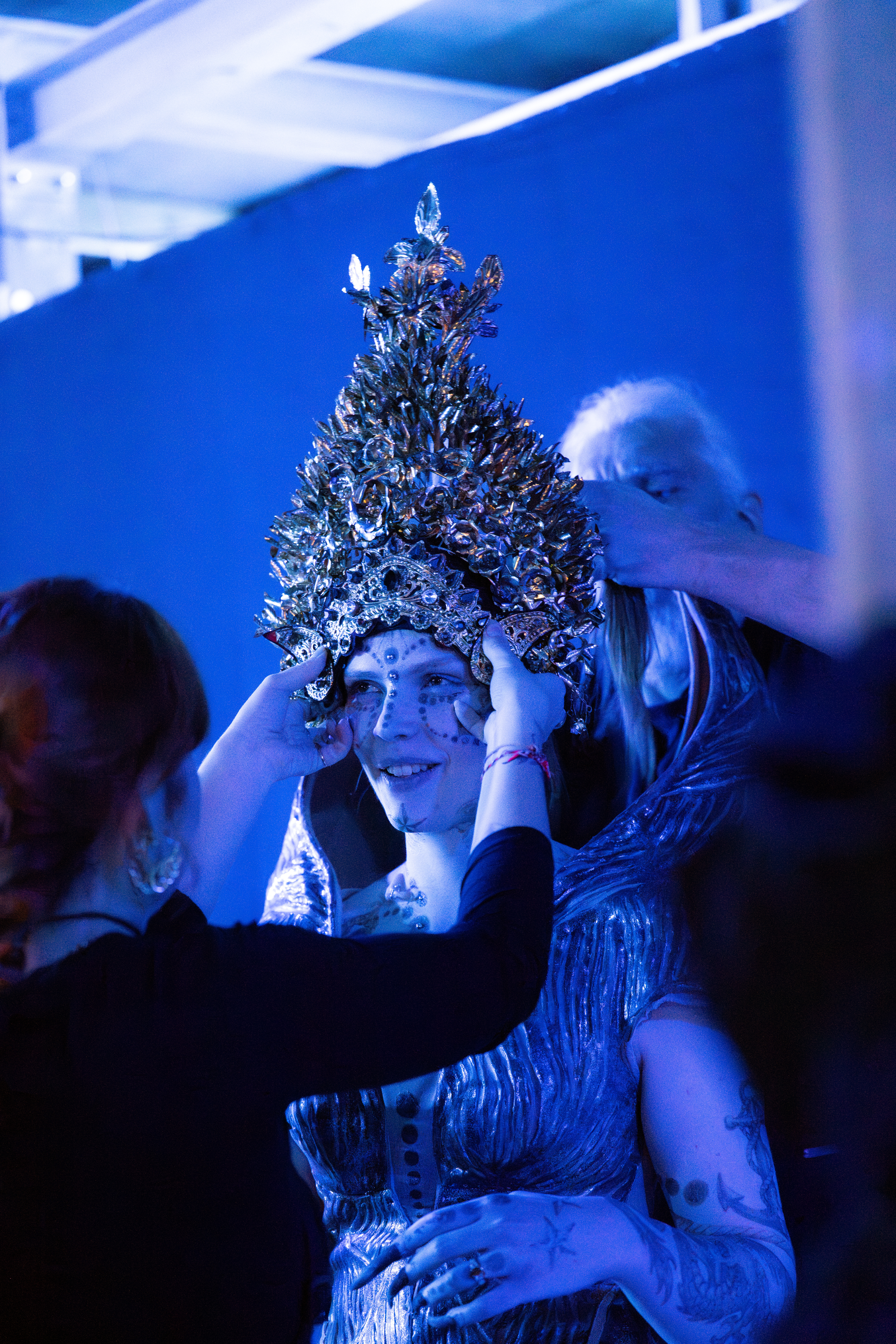 Eine Person mit einem verzierten, silbernen Kopfschmuck und einem schimmernden Outfit wird von zwei Personen unter blauem Licht unterstützt, die den Kopfschmuck und das Kostüm anpassen.
