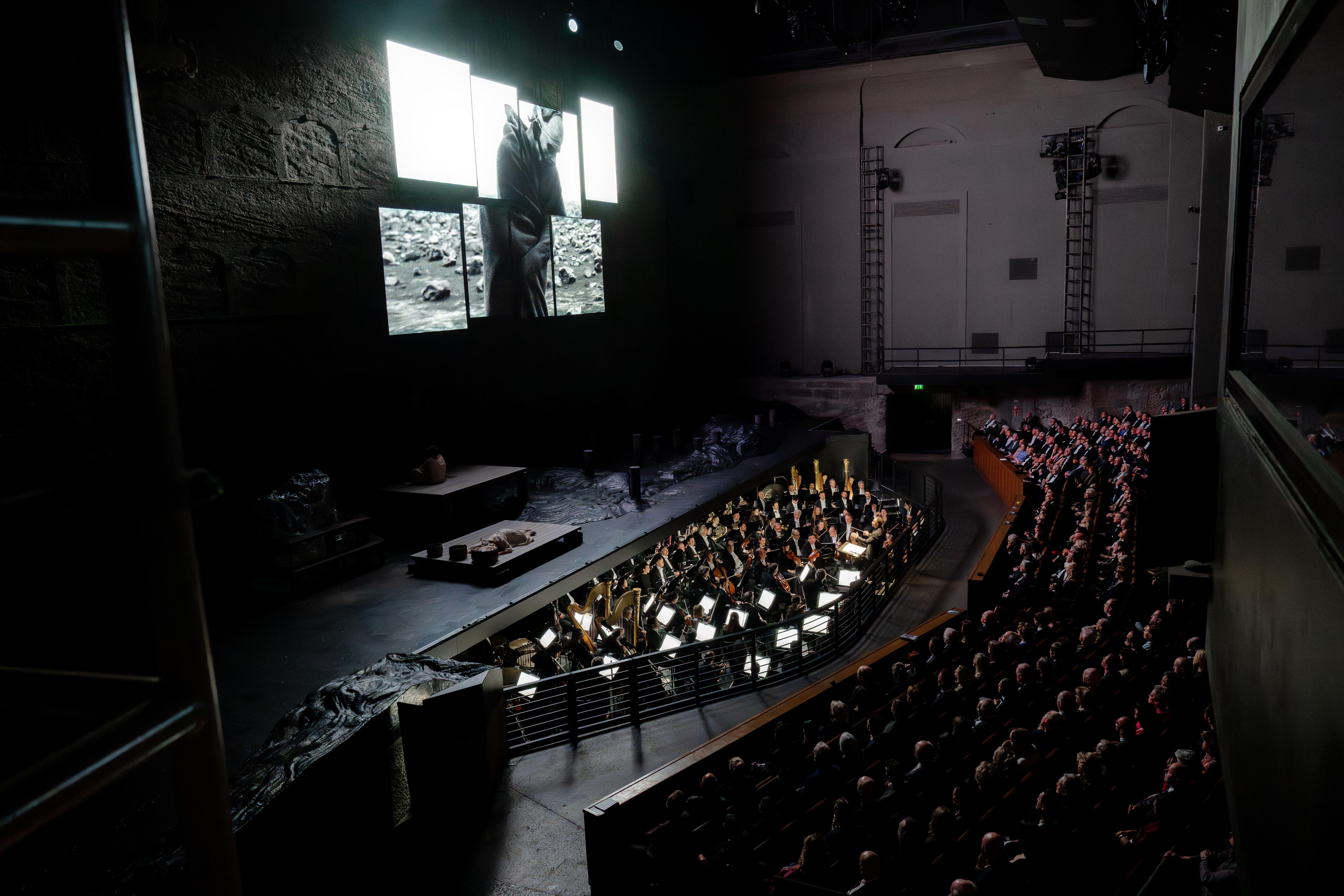 Die Berliner Philharmoniker spielen im Orchestergraben vor vollem Haus, während über den großen Bildschirmen Schwarz-Weiß-Bilder in einer dunklen, dramatischen Theaterkulisse gezeigt werden.