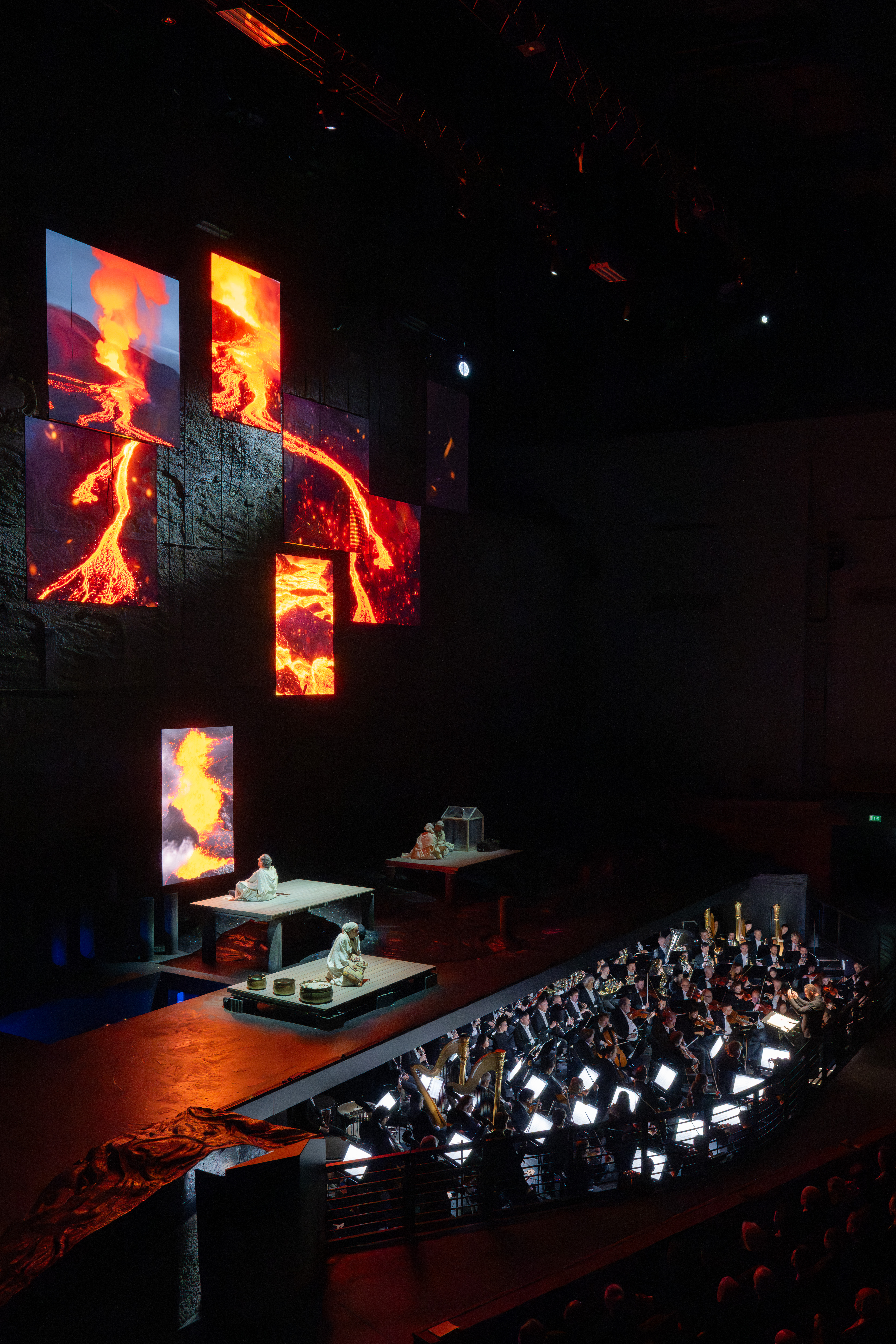 Eine Bühne mit Darstellern befindet sich über einem Orchestergraben, während große vertikale Bildschirme lebendige Bilder von fließender Lava zeigen, die eine dramatische Kulisse in dem schwach beleuchteten Theater bilden.