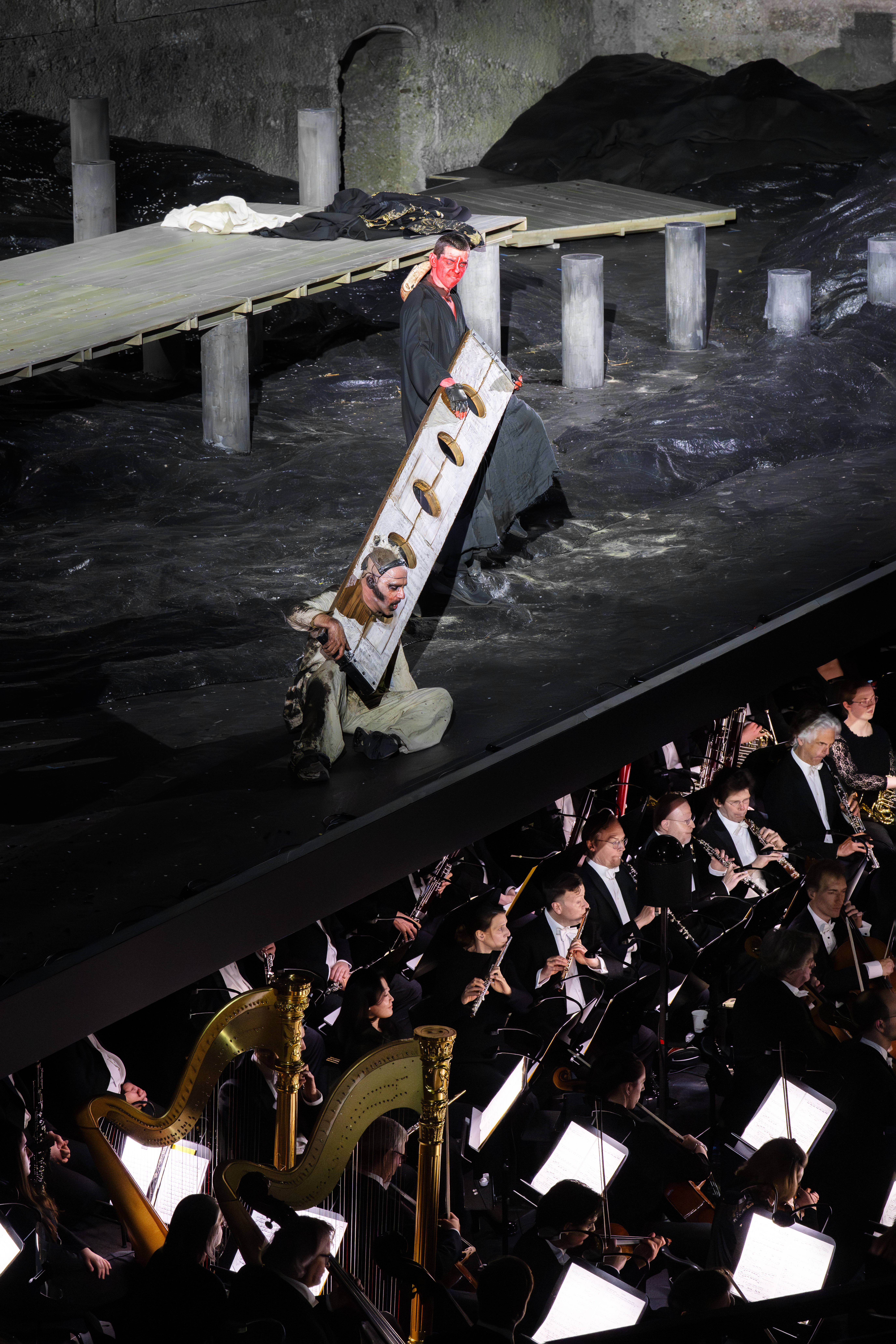 Ein Bühnenkünstler mit Maske steht auf einem schwarzen Bühnenbild mit einem Holzsteg. Unten spielt ein Orchester mit Harfen und Musikern Musik.