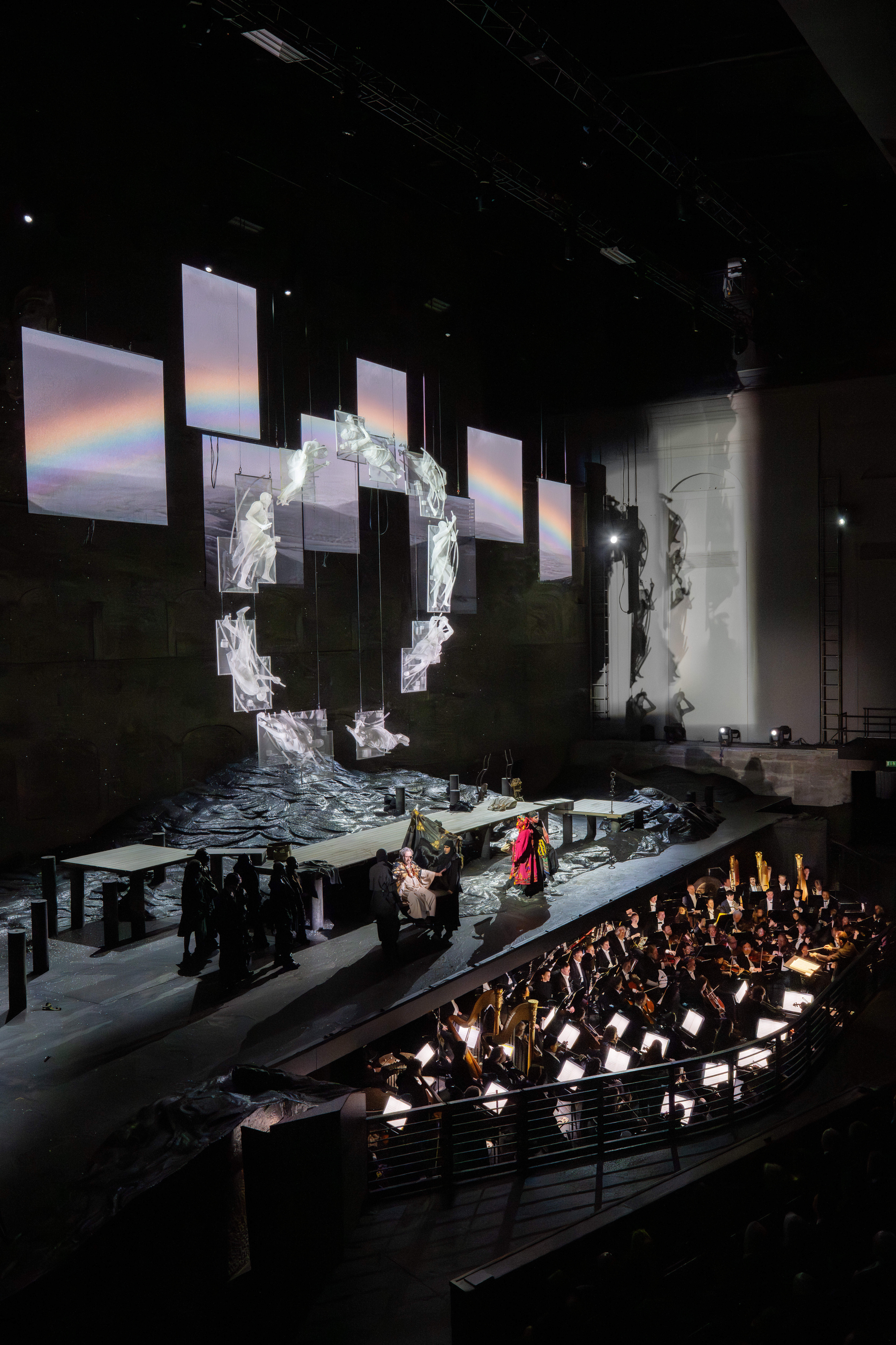 Ein Orchester spielt unter einem dramatischen Bühnenbild mit digitalen Bildschirmen, felsigem Gelände und Darstellern in Kostümen bei theatralischer Beleuchtung in einem großen Theater.