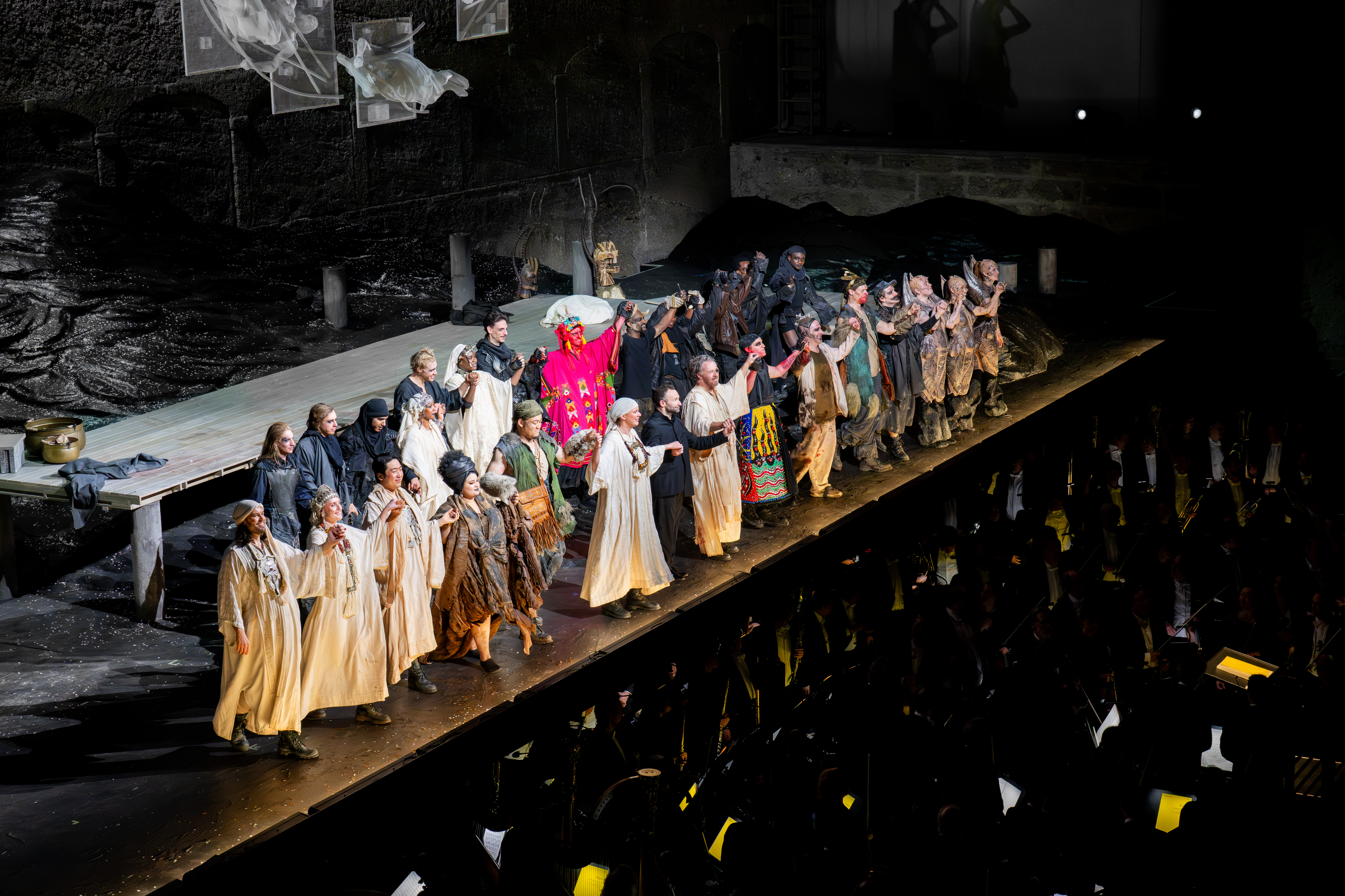 Kirill Petrenko sowie eine große Gruppe von Darstellern in Kostümen stehen auf der Bühne, hält sich an den Händen und verbeugen sich vor dem applaudierenden Publikum nach einer Theateraufführung, während im Hintergrund Kulissen und dramatische Beleuchtung zu sehen sind.