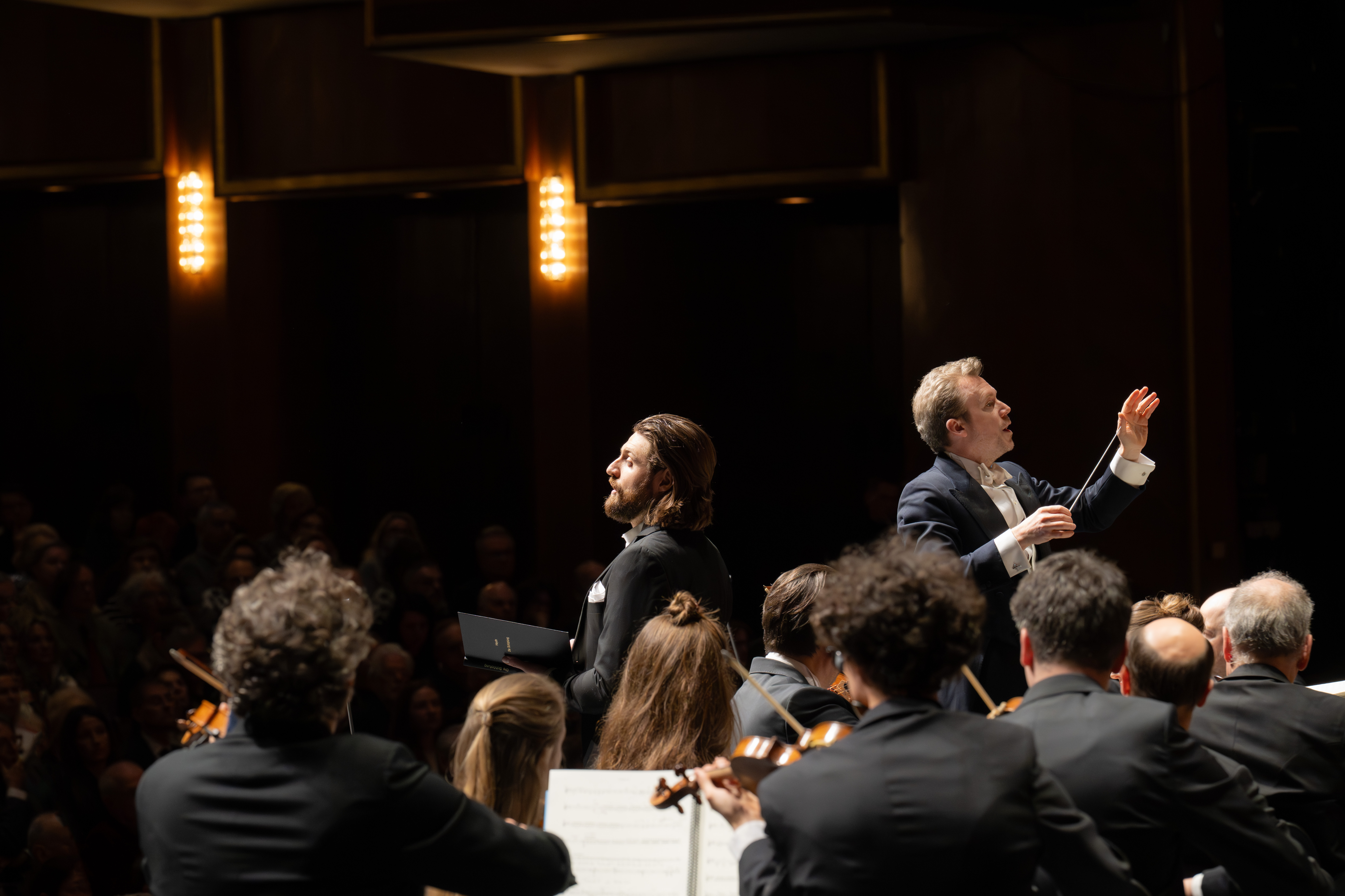 Ein Dirigent leitet ein Orchester auf der Bühne, auf der Musiker Streichinstrumente spielen und ein Publikum in einem schwach beleuchteten Konzertsaal zusieht. Ein Sänger steht mit einer Notenmappe da.