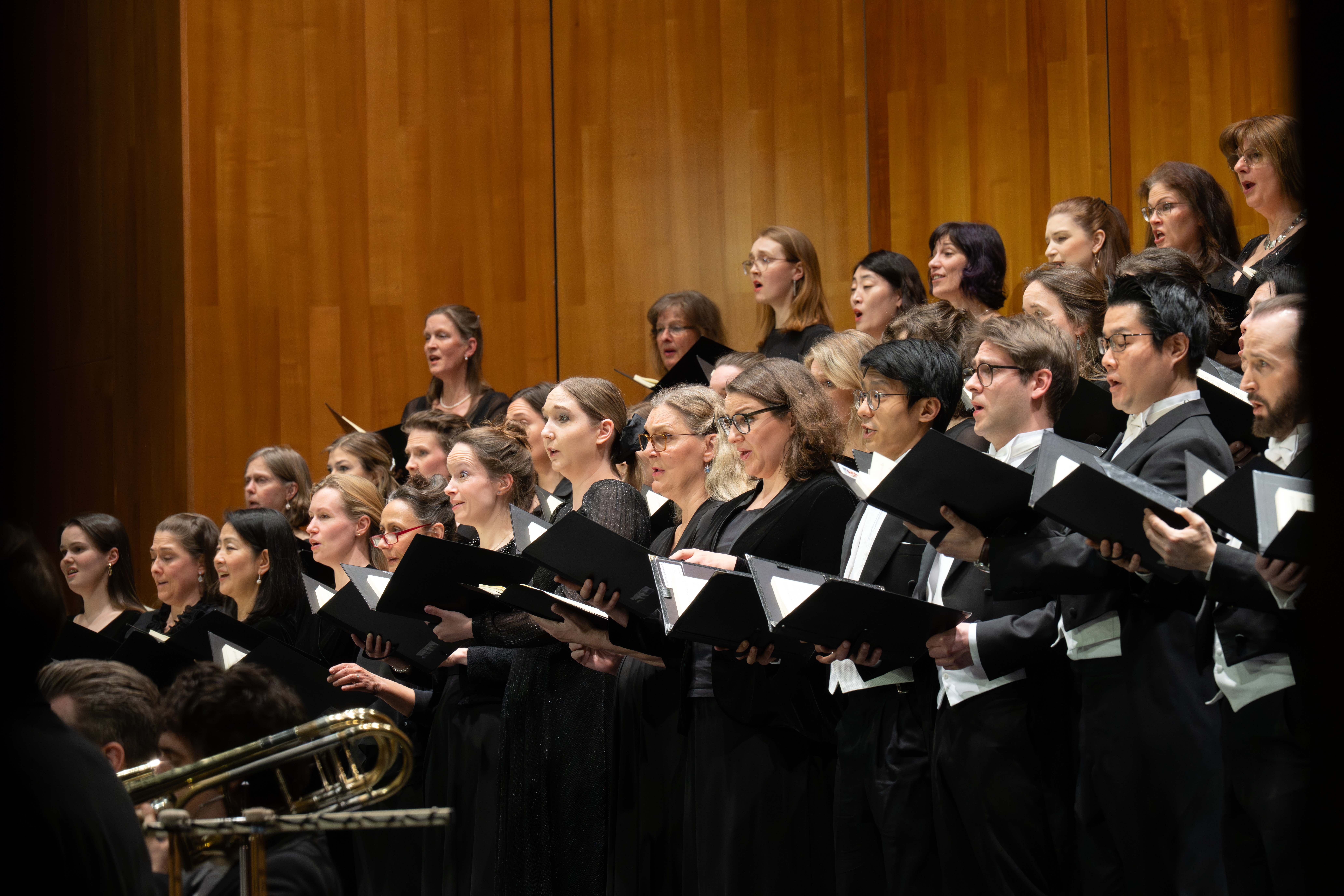 Ein Chor von Männern und Frauen in schwarzer Kleidung singt auf der Bühne, in der Hand offene Notenmappen, im Hintergrund eine Holzverkleidung.
