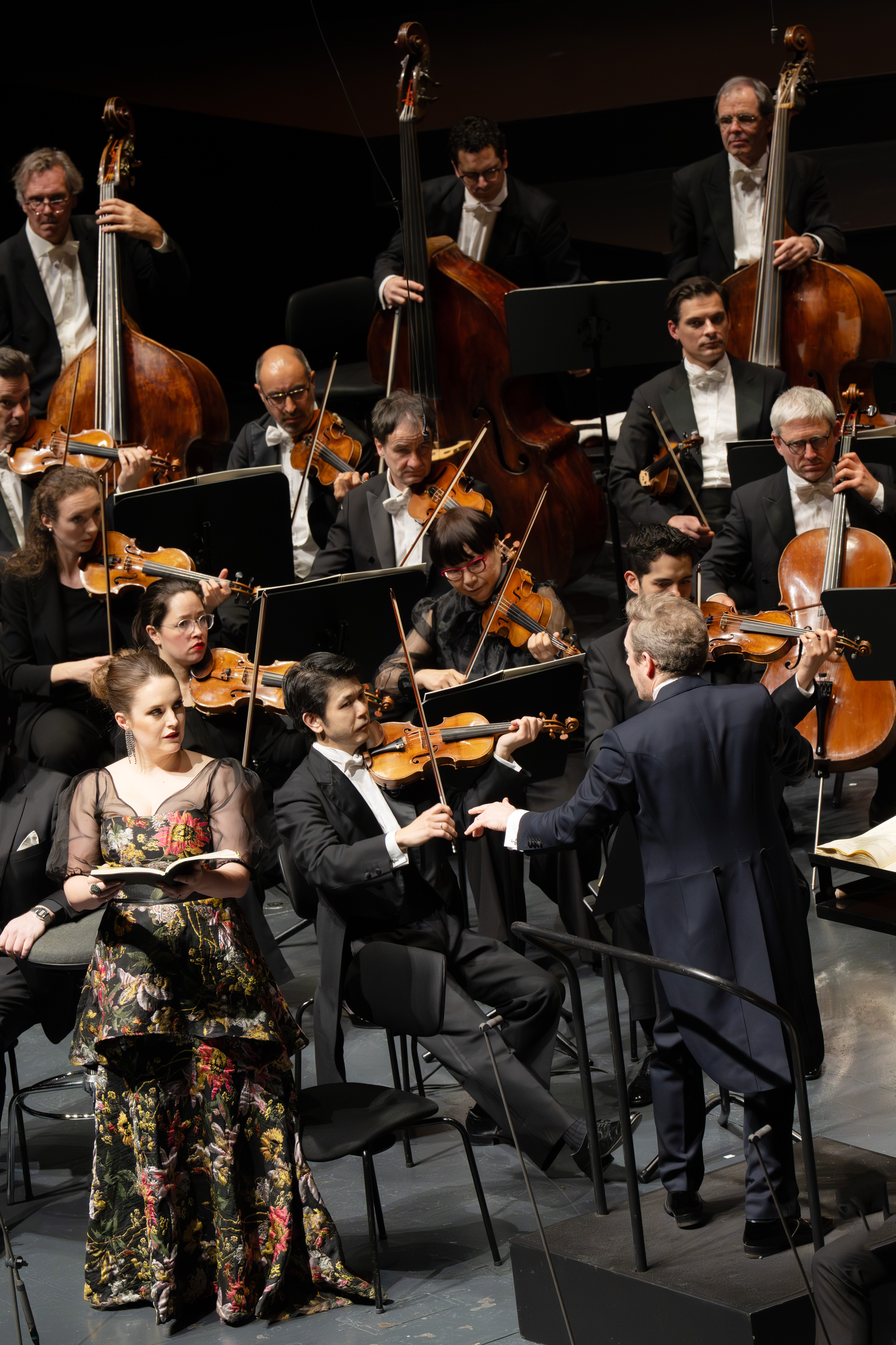 Daniel Harding leitet ein Orchester in formeller Kleidung. Eine Solistin in einem geblümten Kleid steht an der Spitze, hält Noten in der Hand und singt. Die Musiker spielen Geigen, Celli und Kontrabässe.