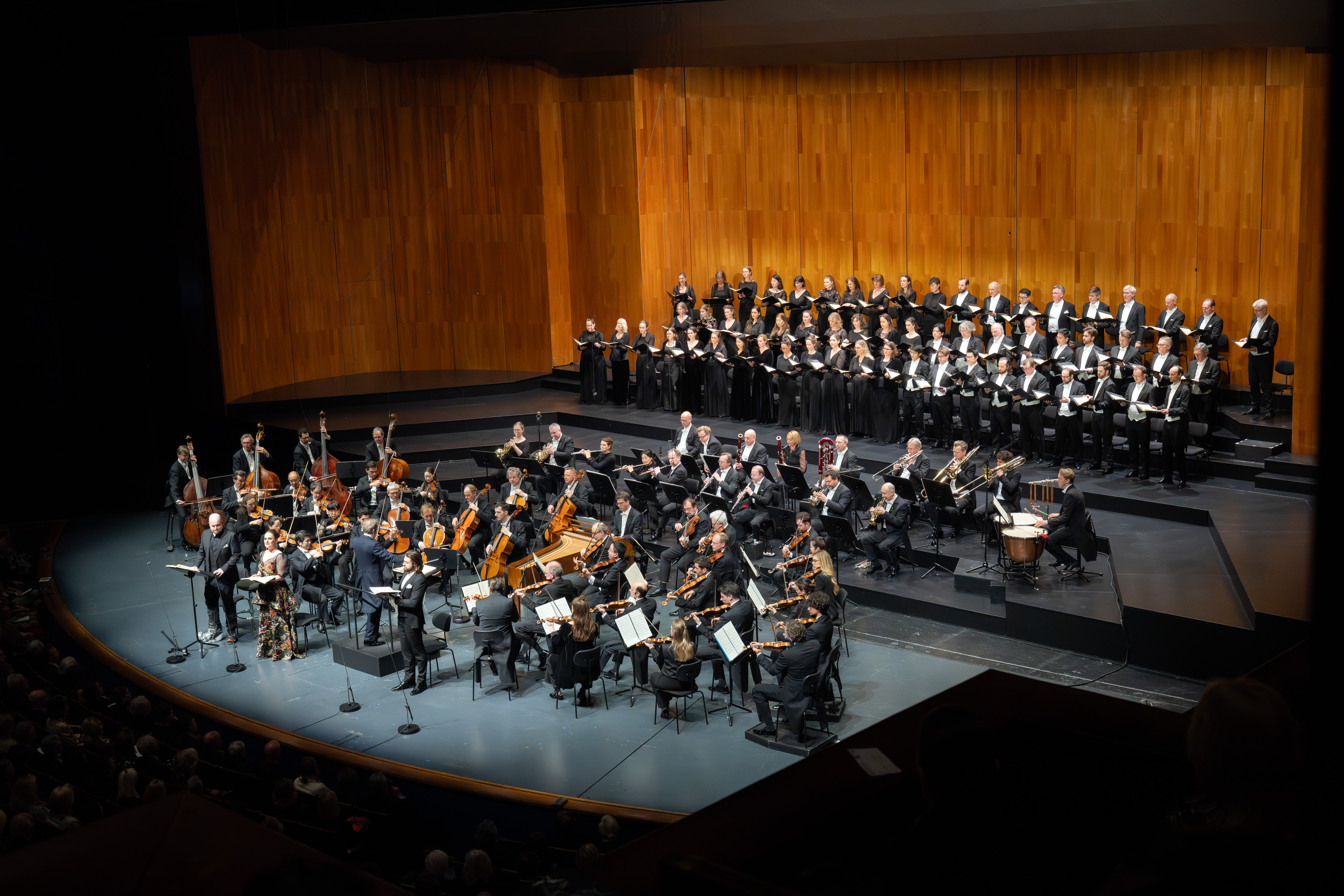 In einem Konzertsaal mit hölzernem Hintergrund und einem Publikum im Vordergrund tritt auf der Bühne ein großes Orchester auf, hinter dem ein großer Chor steht, alle in Schwarz gekleidet.