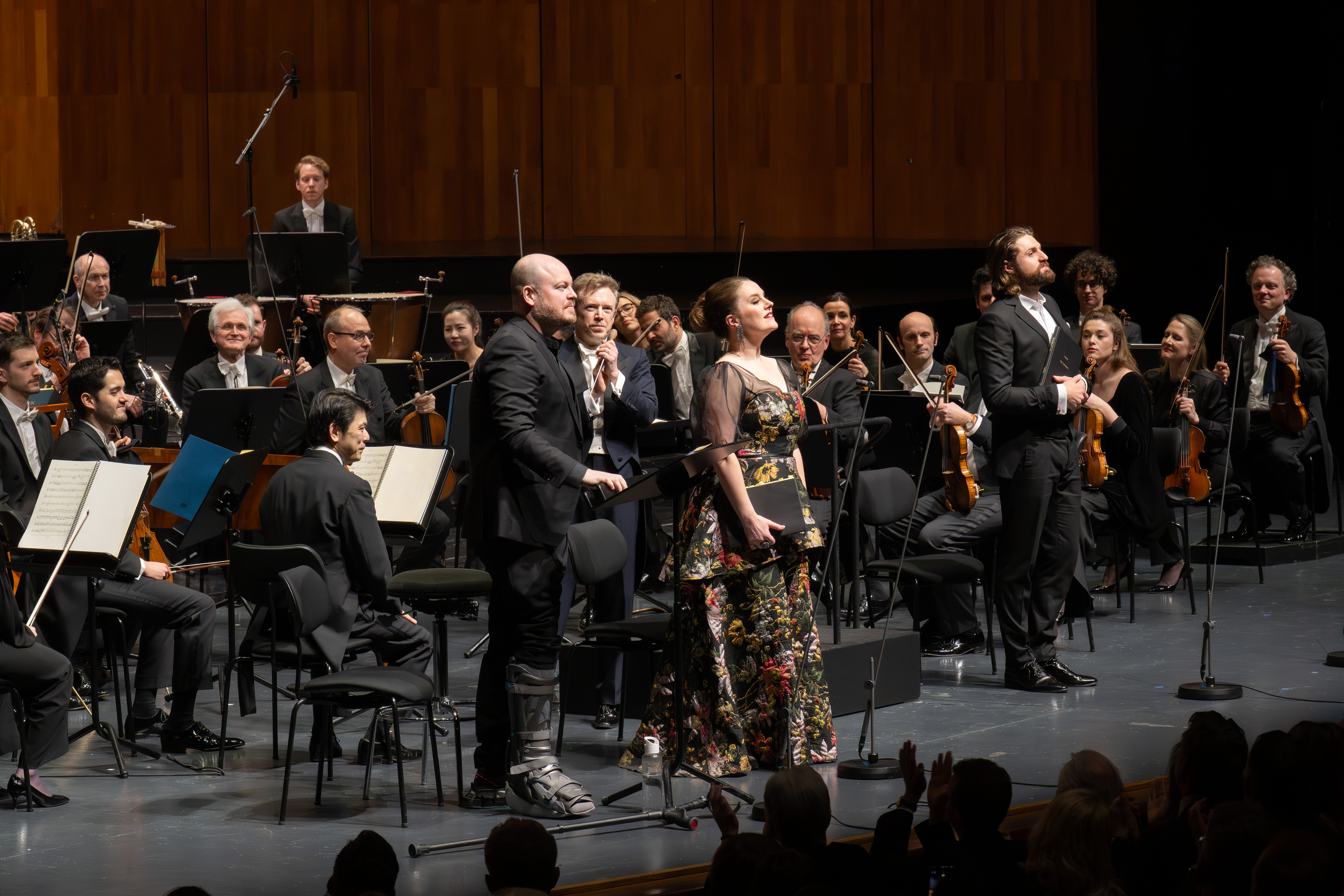 Eine Frau in einem geblümten Kleid steht auf der Bühne und hält Blumen, flankiert von zwei Männern in Anzügen, während hinter ihnen ein Orchester sitzt. Im Vordergrund ist das Publikum zu sehen, das applaudiert.