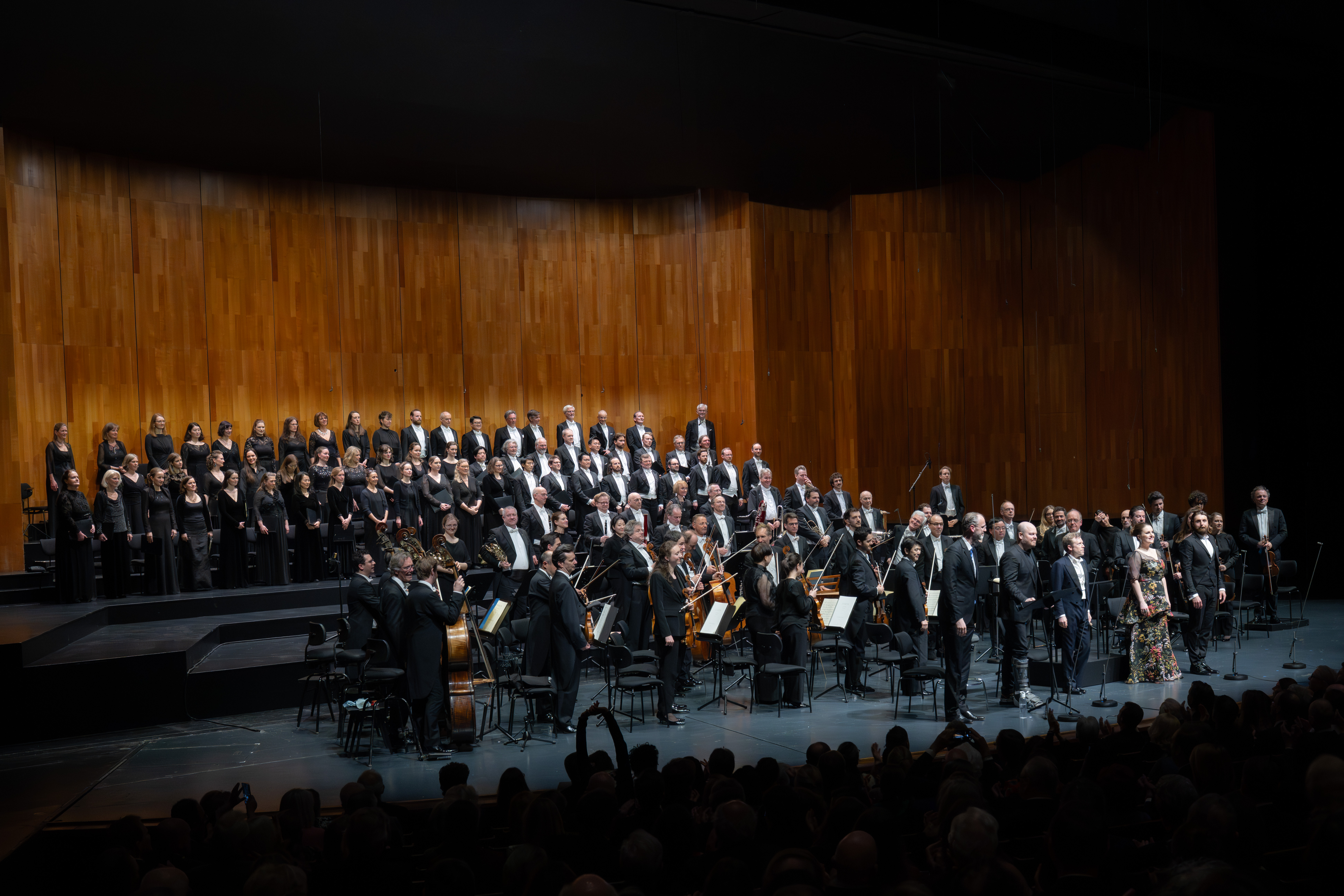 Ein großes Orchester und ein Chor, alle in formeller schwarzer Kleidung, treten auf einer hell erleuchteten Holzbühne auf, während das Publikum im abgedunkelten Vordergrund zusieht.
