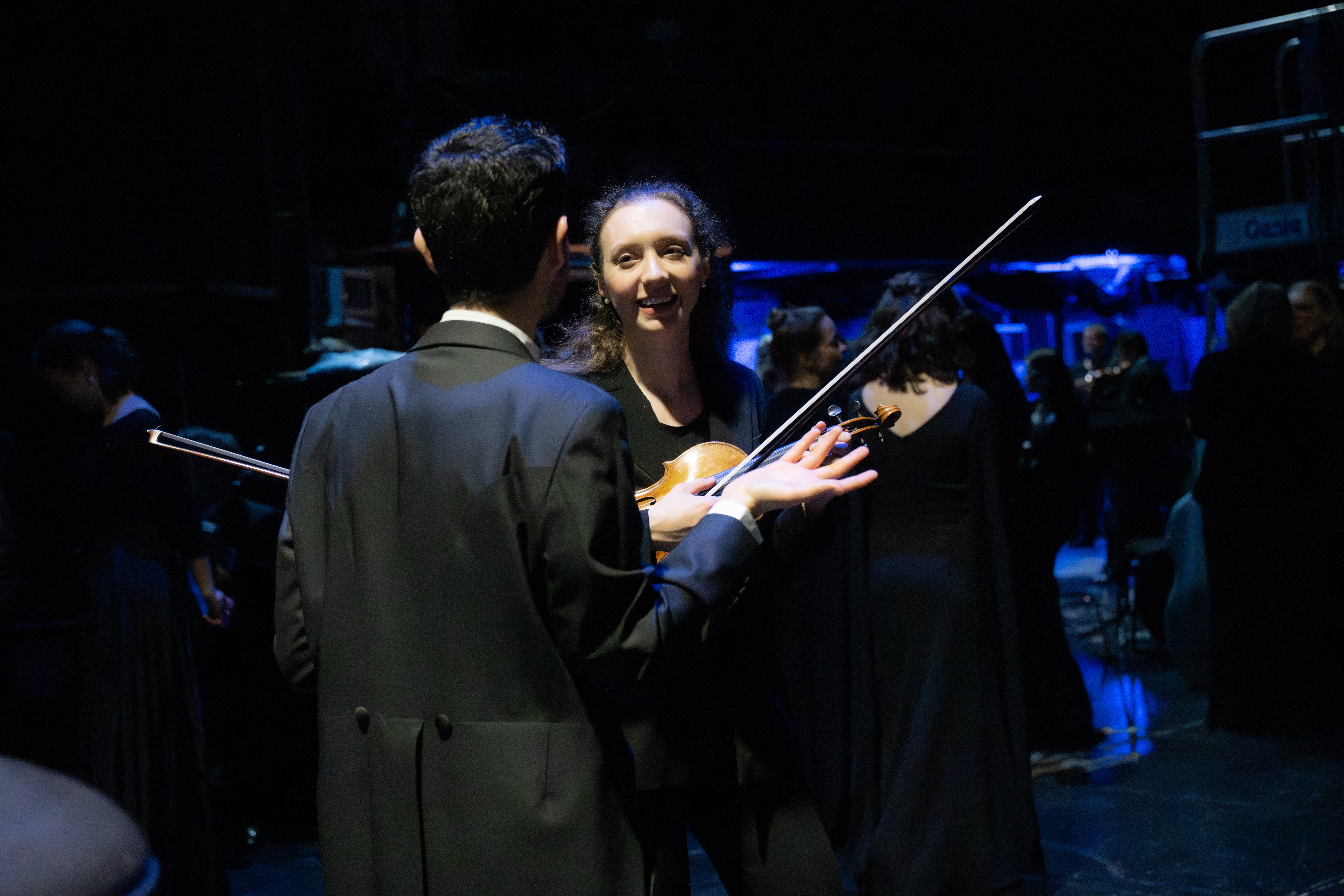 Ein Orchestermusiker und eine Orchestermusikerin in formeller schwarzer Kleidung unterhalten sich hinter der Bühne, einer hält eine Geige und lächelt, während andere Musiker im schwach beleuchteten Hintergrund verschwimmen.
