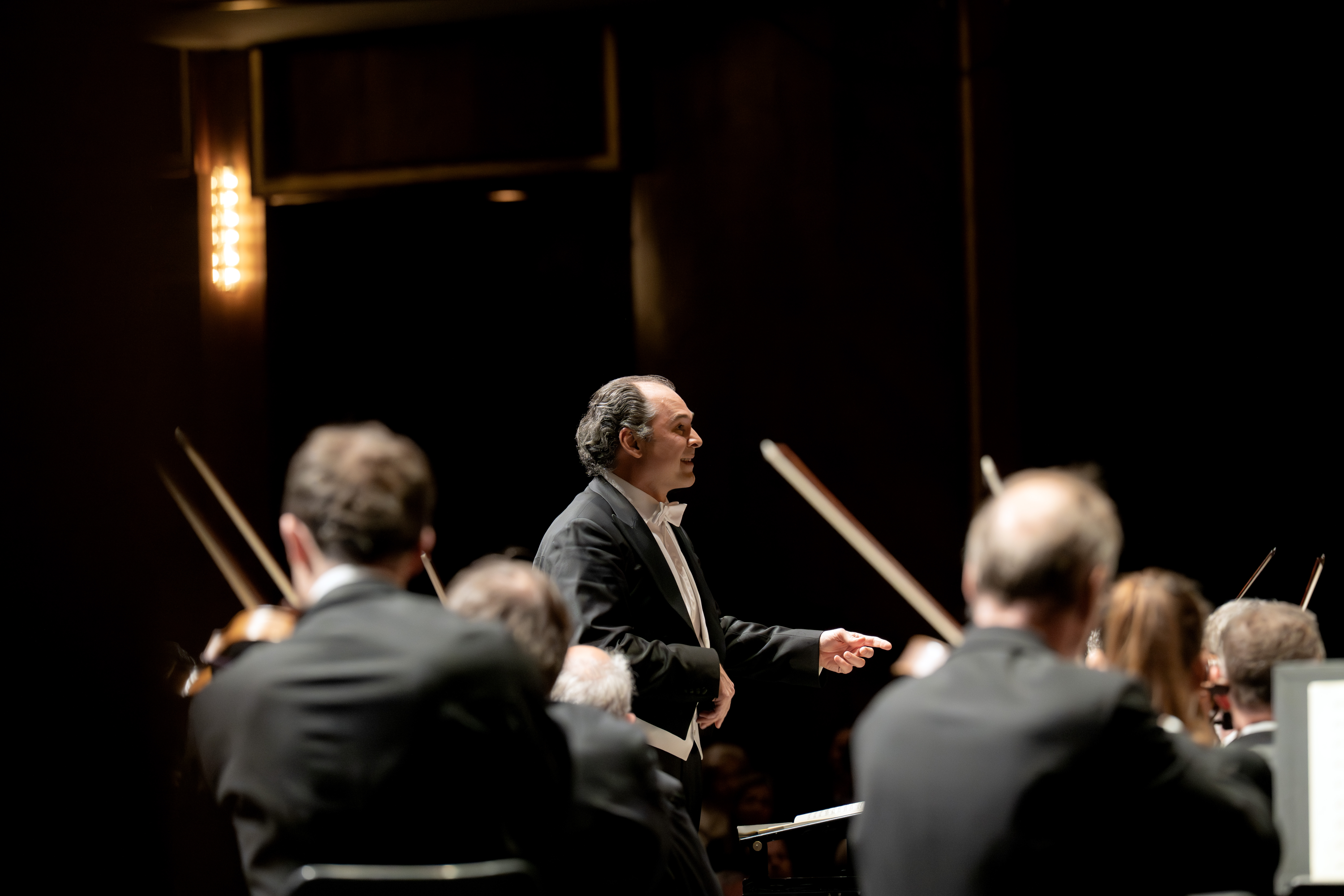 Tugan Sokhiev steht vor einem Orchester und führt die Musiker mit erhobenen Armen. Musiker in dunkler, förmlicher Kleidung spielen im Schein der Bühnenbeleuchtung auf Streichinstrumenten. Der Hintergrund ist dunkel und unscharf.